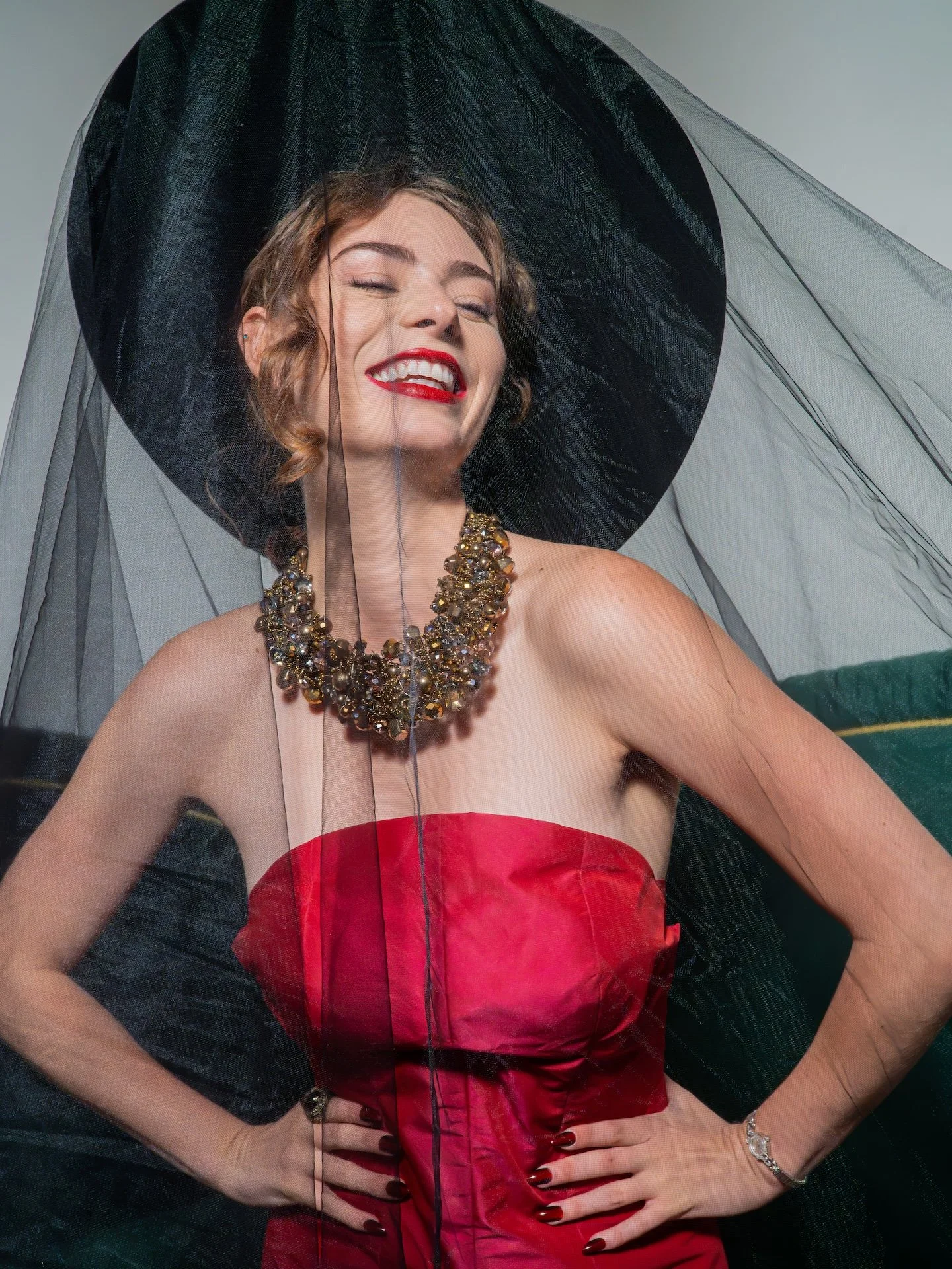 A woman smiling with eyes closed, wearing a large black hat with sheer black veil, a strapless red dress, a chunky multi-colored necklace, and jewelry on her fingers and wrist.