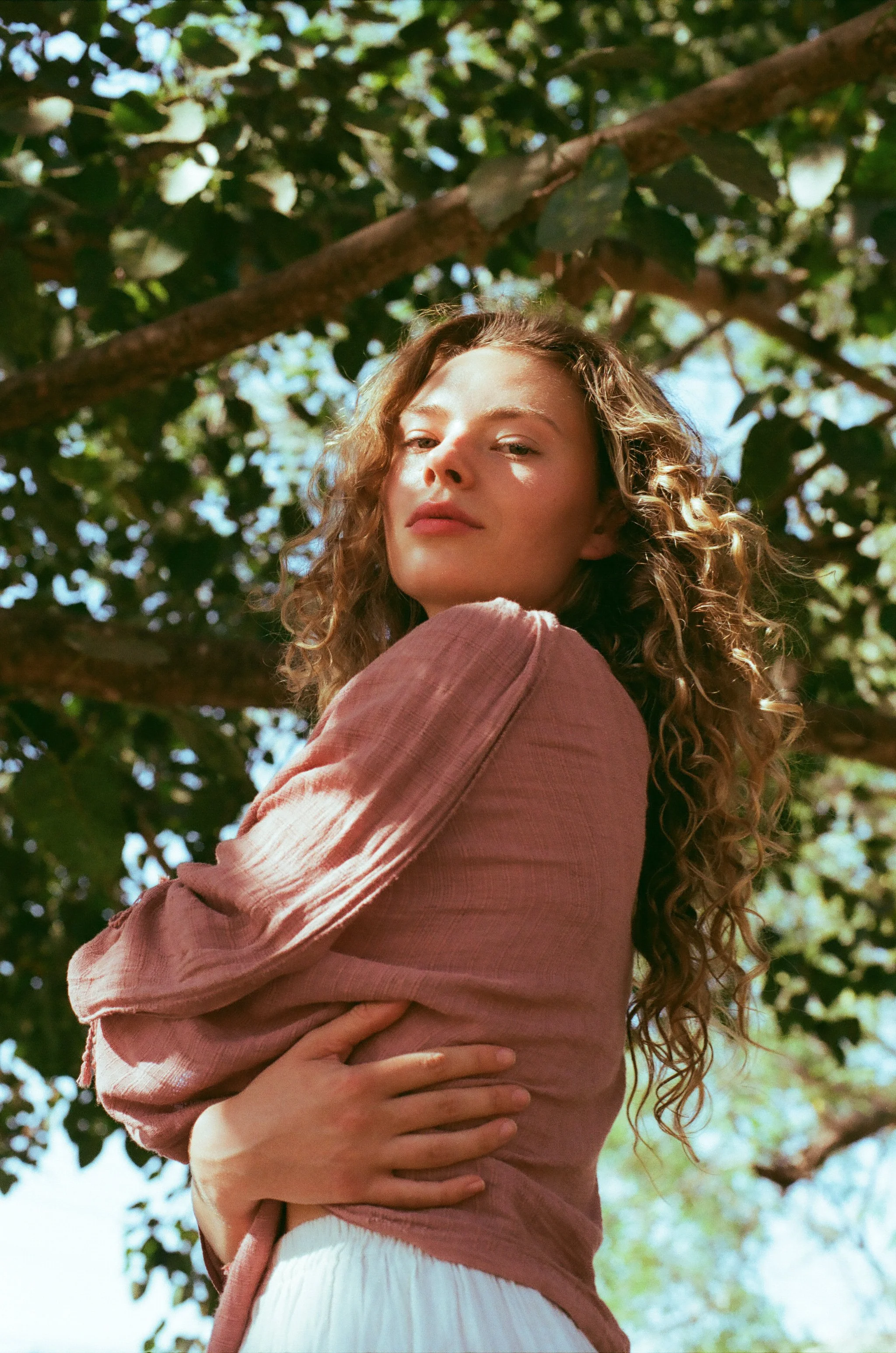 A woman with curly hair standing outdoors with her arms crossed against her chest. She is wearing a pinkish blouse and white pants, with sunlight filtering through green leaves in the background.
