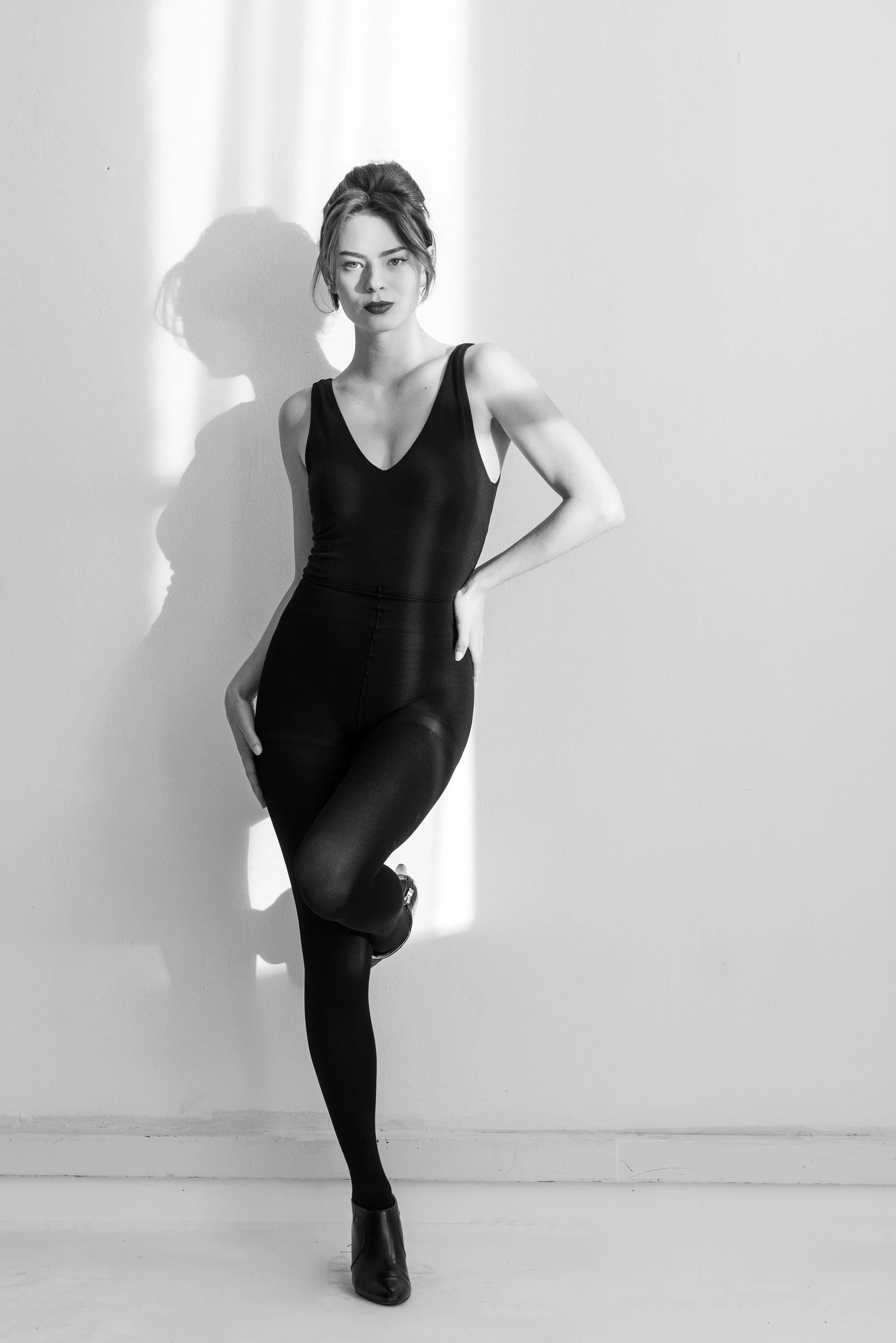 Black and white photo of a woman in a sleeveless dress and high heel boots, standing against a plain wall, with a focused expression, one arm on her hip, and one foot crossed in front of the other, casting a shadow on the wall.