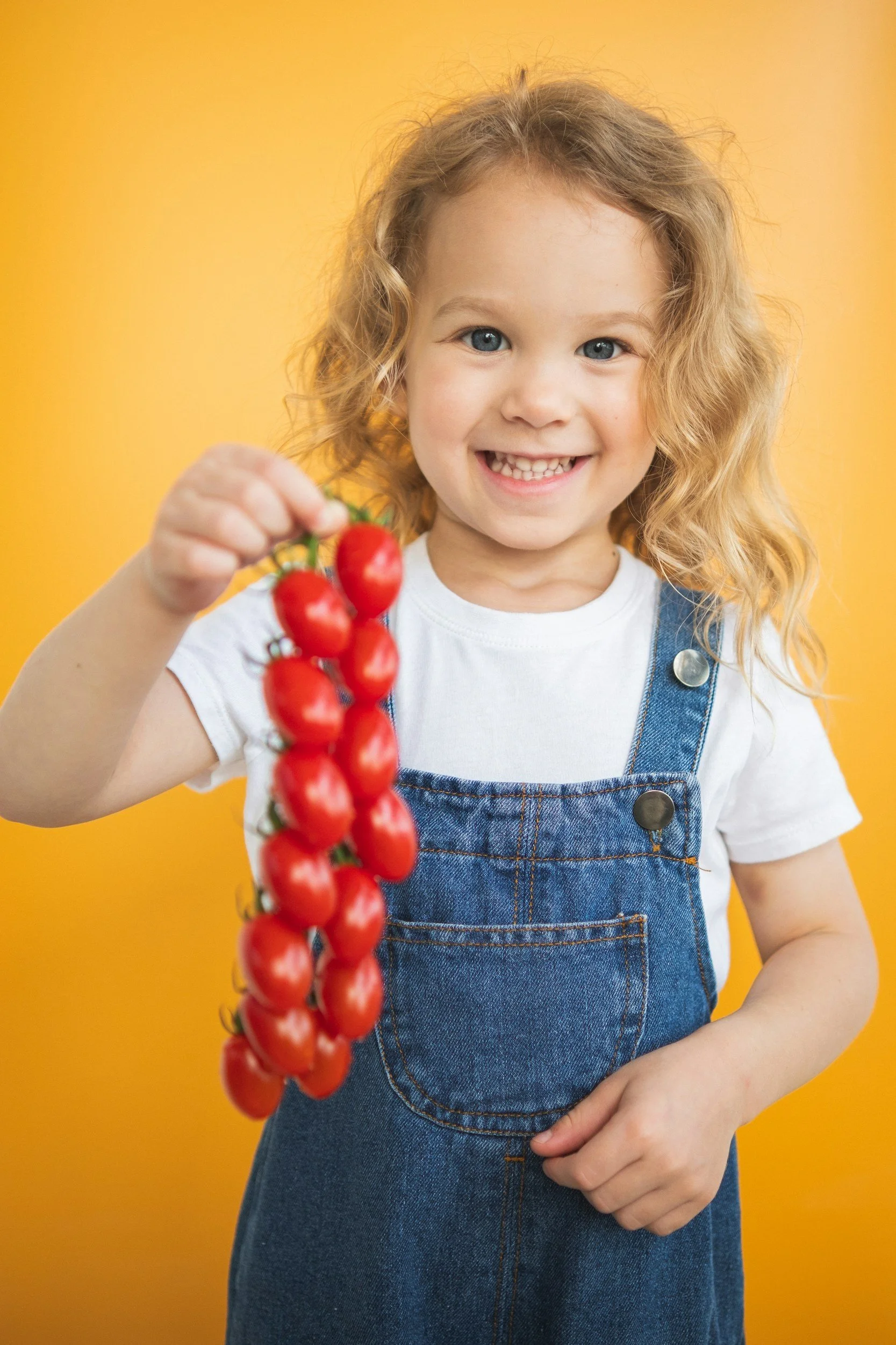 A young girl with curly blonde hair and blue eyes smiling and holding a bunch of red cherry tomatoes on a yellow background.