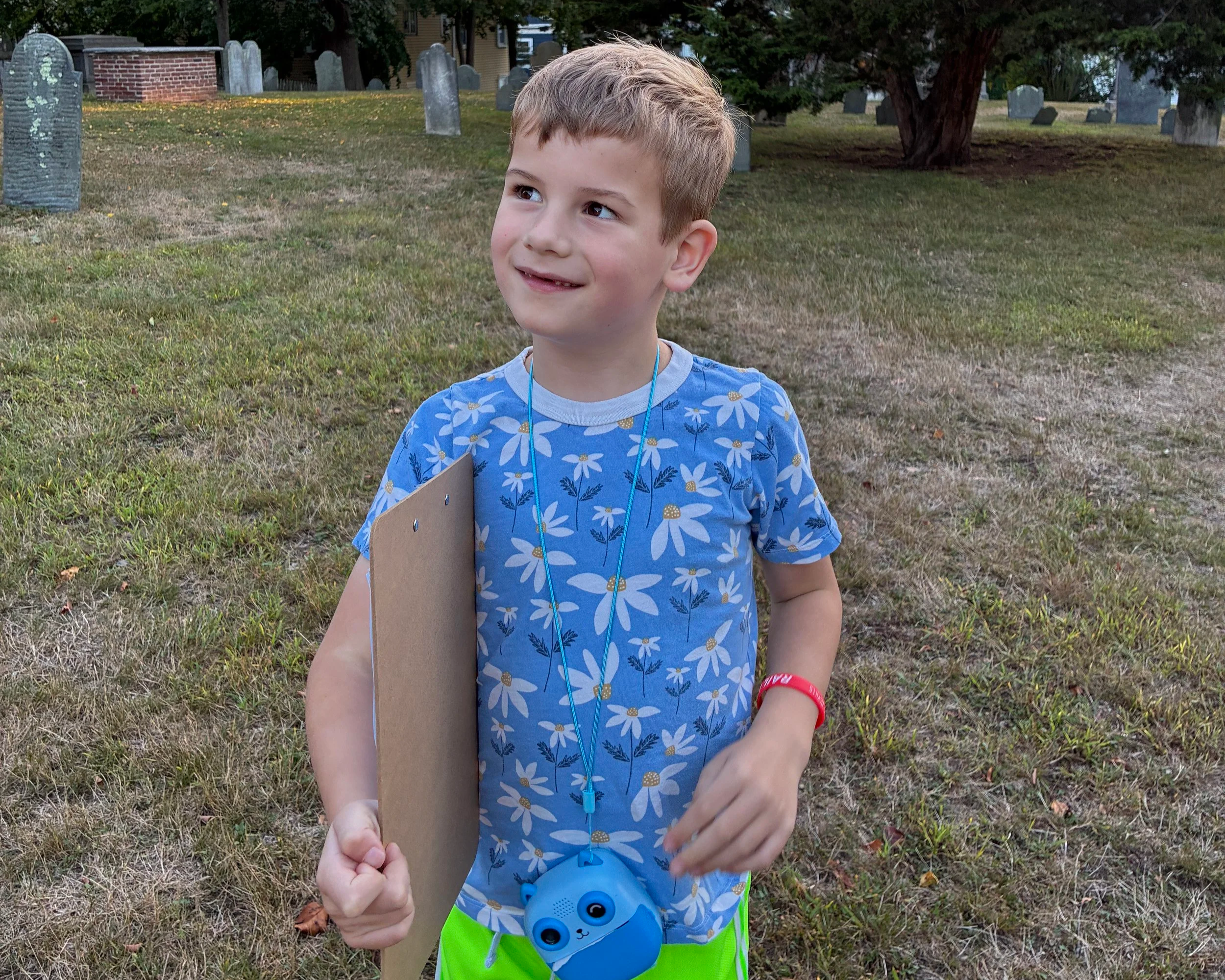 A young boy with short blond hair, wearing a blue T-shirt with white daisy patterns, green shorts, and a red wristband, stands in a grassy area with gravestones visible in the background.