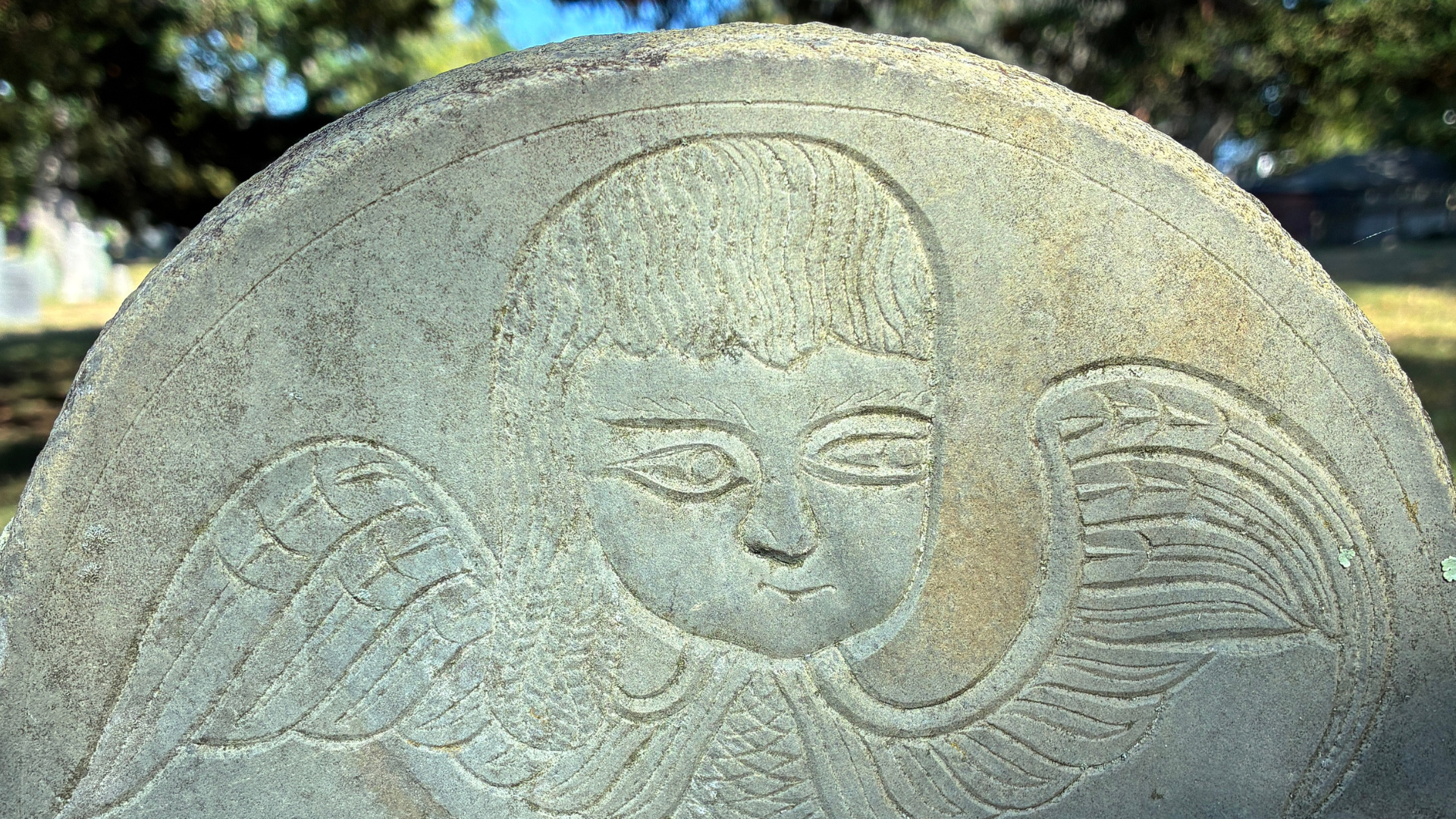 Close-up of a carved stone featuring a stylized face with closed eyes and flowing hair, set outdoors with trees and a bright sky in the background.