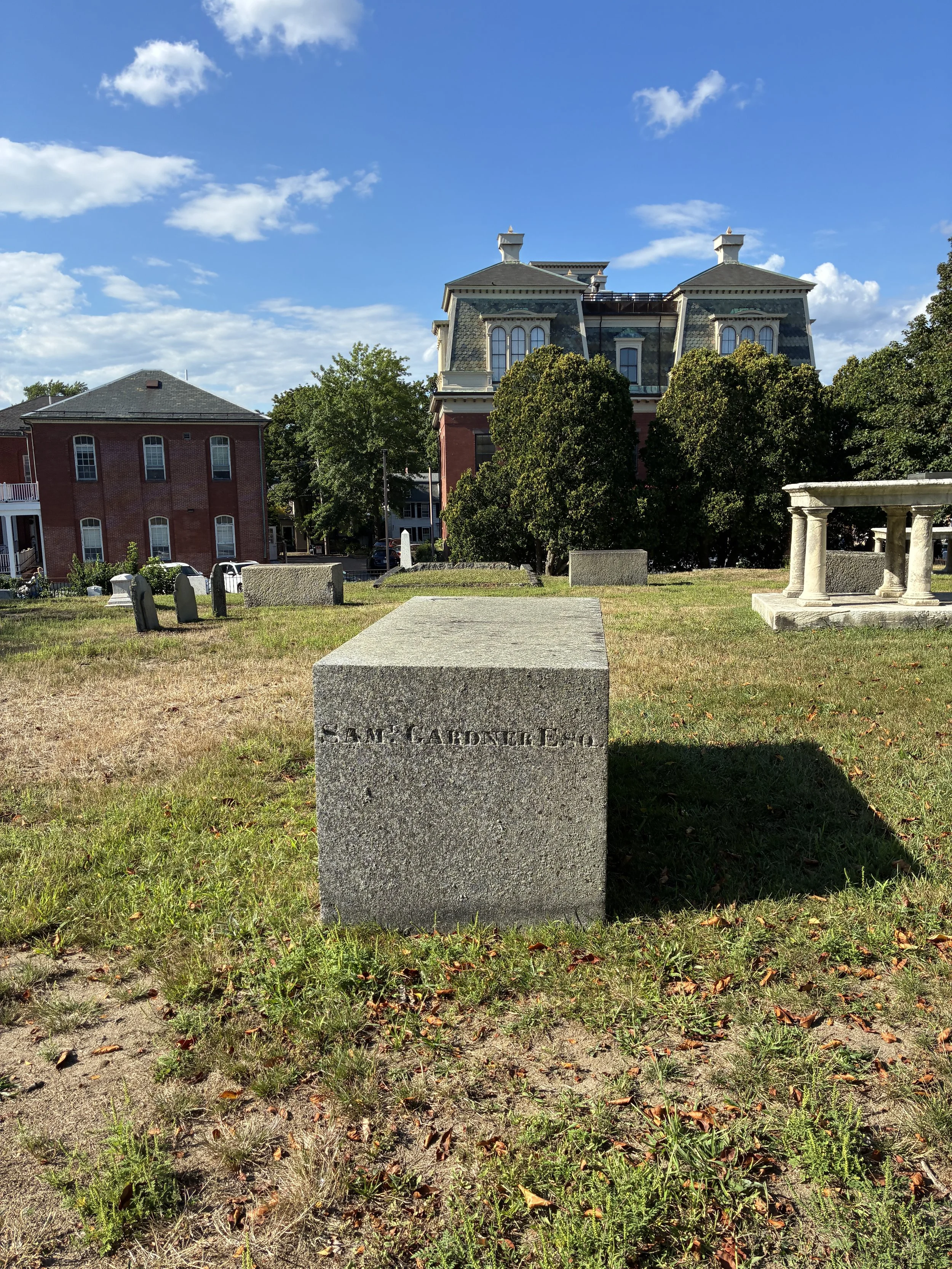 Gravestone in a grassy area with trees and historic buildings in the background on a sunny day.