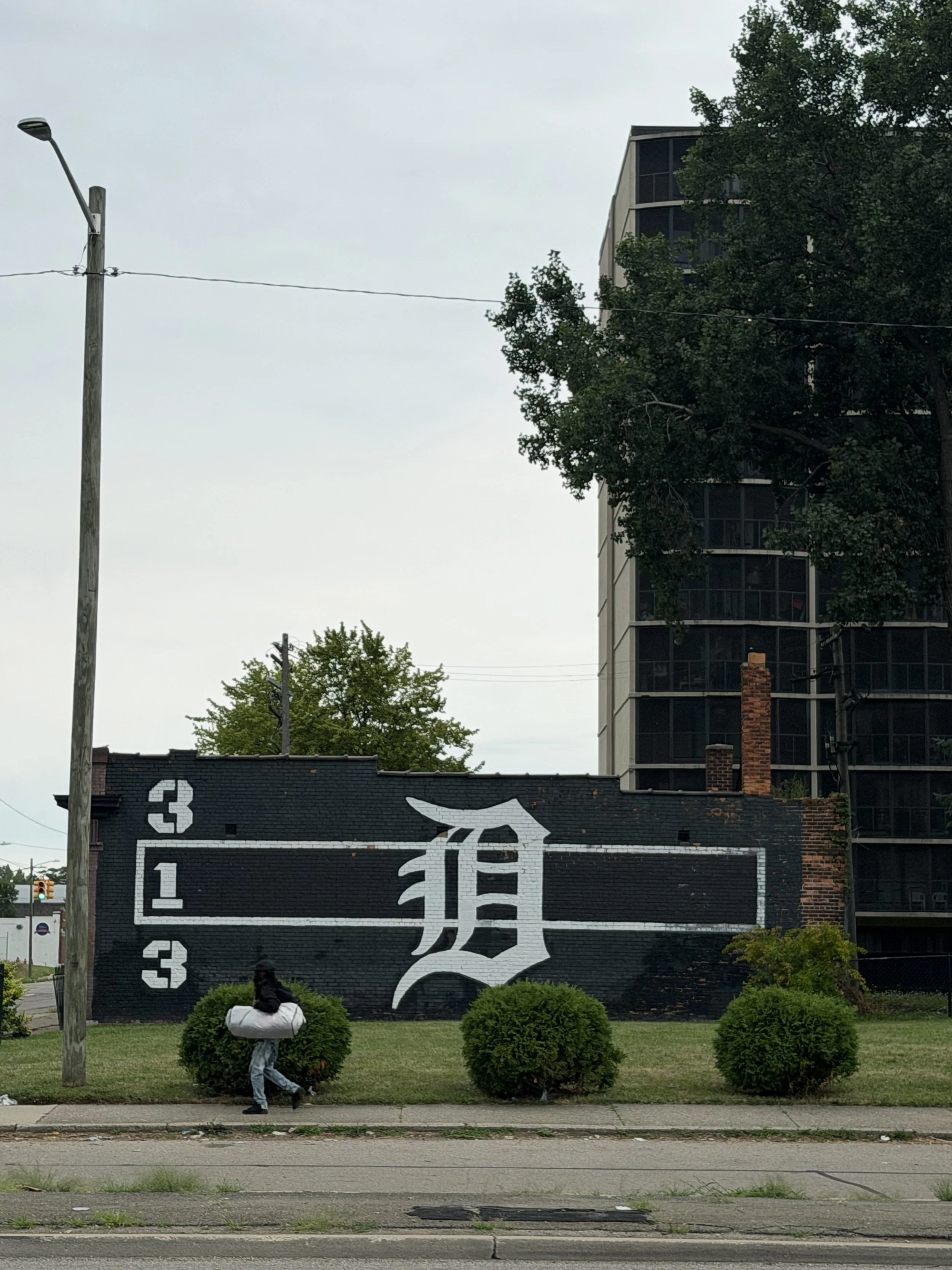 A mural painted on a brick wall displaying a large stylized Chinese character in white with black outlines, alongside vertical white numbers reading 3, 1, 2, and 3, and a smaller white emblem resembling a bat or wings. The mural is surrounded by four neatly trimmed round bushes, with a woman walking by carrying a white bag and wearing a black jacket and light-colored pants. There is a sidewalk in front of the mural and the sky appears overcast.