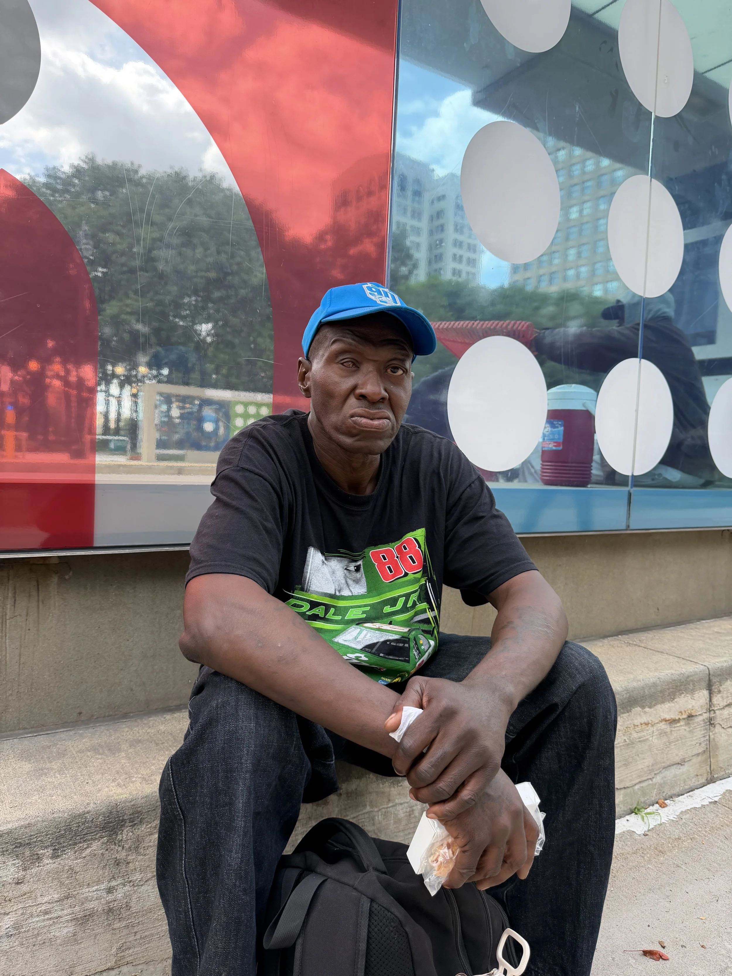 A man wearing a blue cap and a black t-shirt with a racing graphic sits on the sidewalk holding a sandwich. Behind him is a reflective glass wall with white circle patterns, and a cityscape with trees, a building, and a trash can. The man has a serious expression.