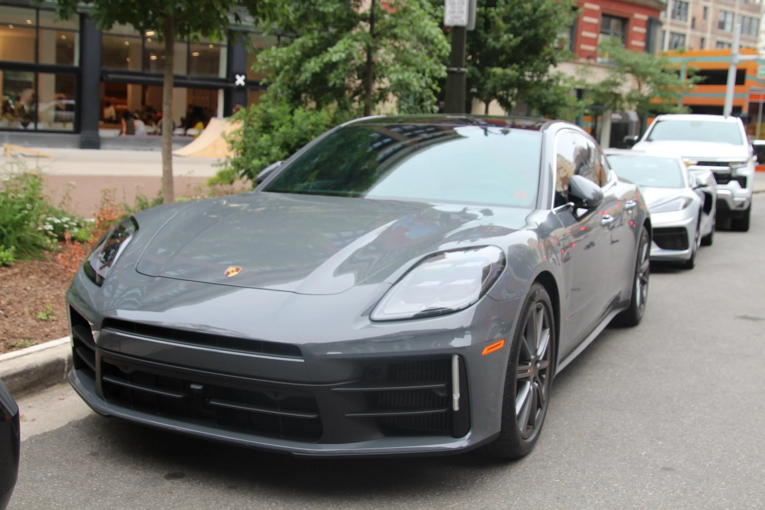 A gray sports car with a Porsche emblem is parked along a city street, with a white SUV parked behind it. In the background, there is a storefront with large windows, some trees, and multi-story buildings.