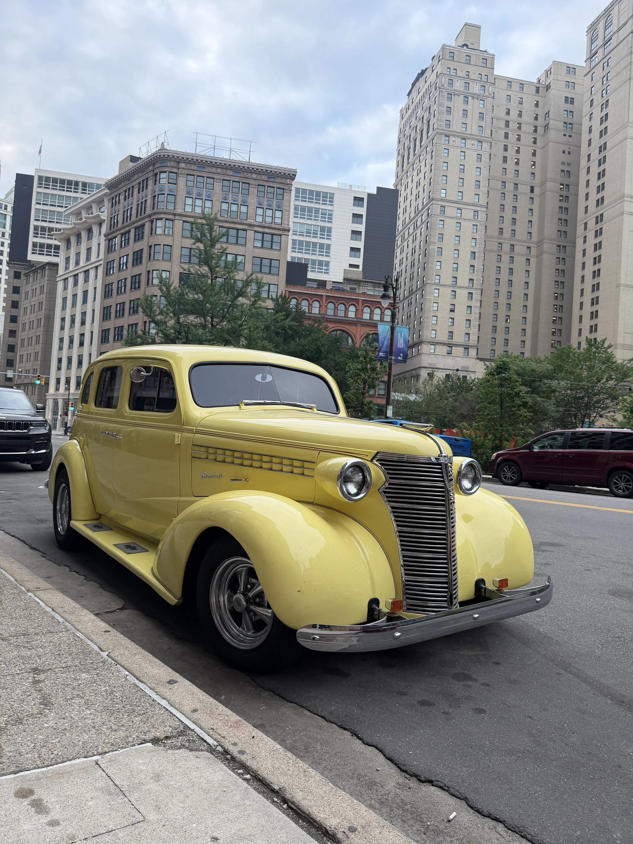 A vintage yellow car parked on the street in front of tall buildings with a cloudy sky overhead.