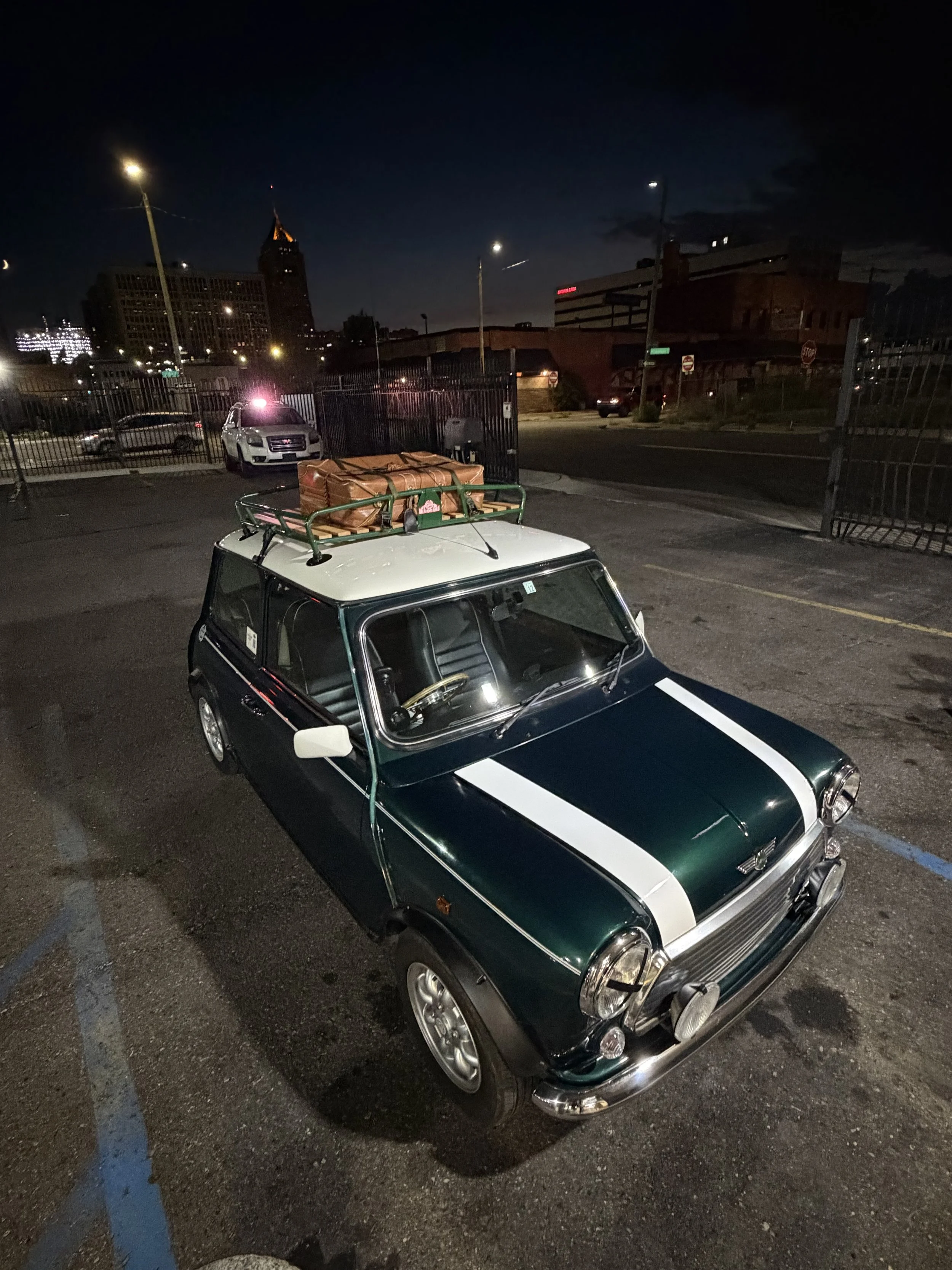 Nighttime city parking lot with a green and white vintage car in foreground, balcony on roof, and city skyline with tall buildings and a clock tower in the background.