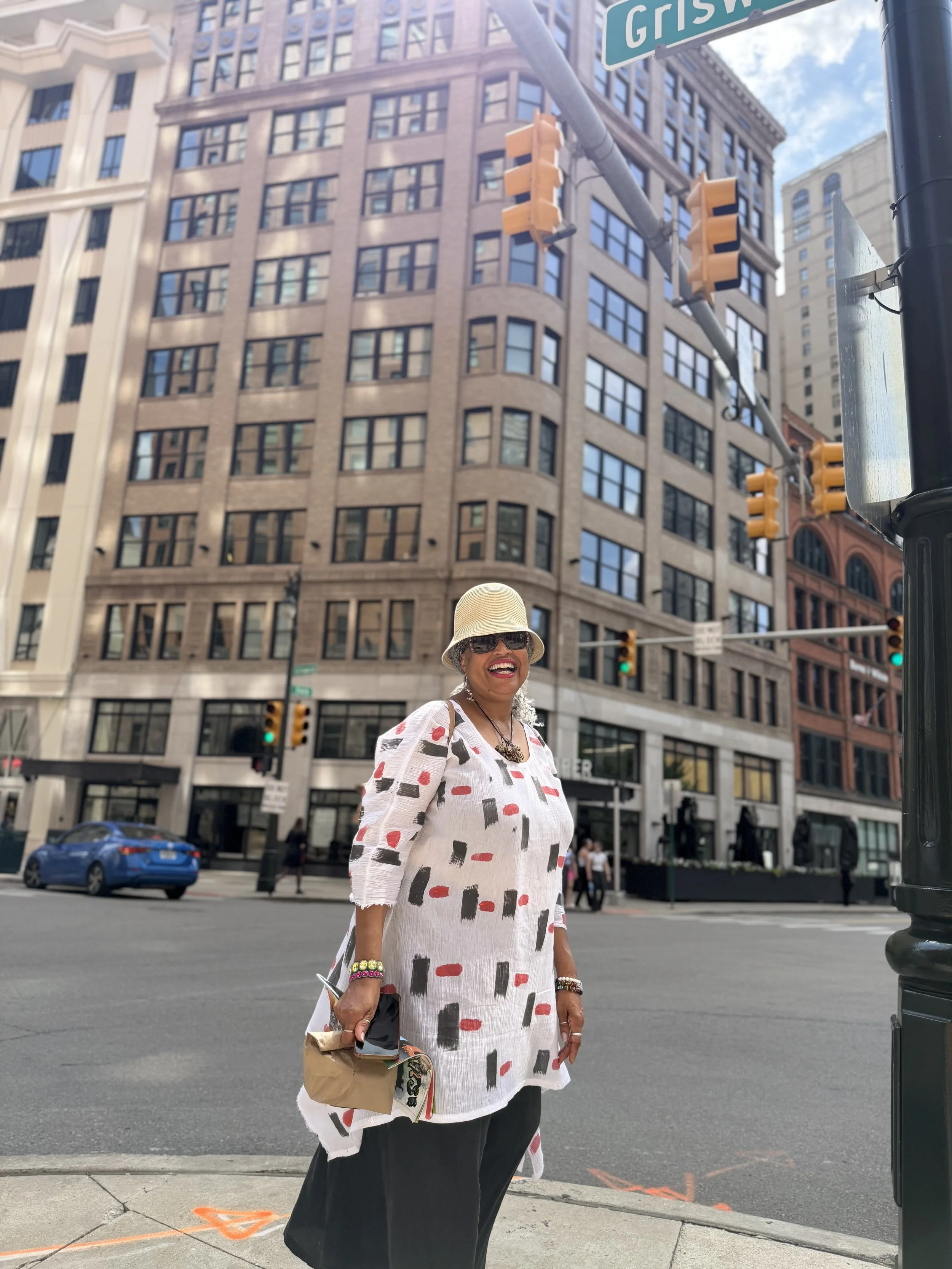A woman smiling and standing on a city street corner with tall buildings in the background. She is wearing a patterned dress, sunglasses, and a hat, and holding a handbag and phone.