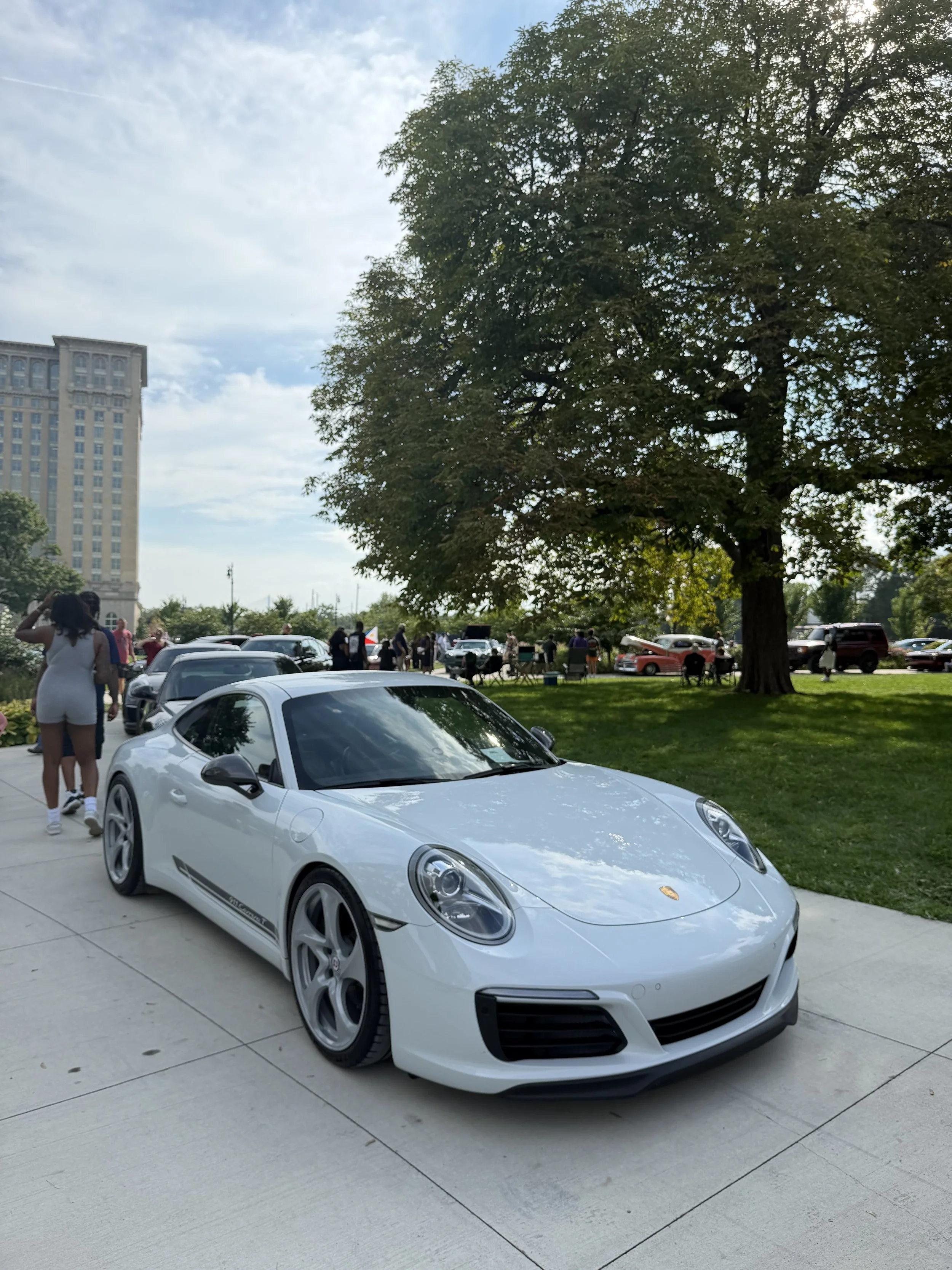 A white Porsche sports car parked on a paved area with people walking and socializing nearby under a large tree. A tall building is visible in the background under a partly cloudy sky.