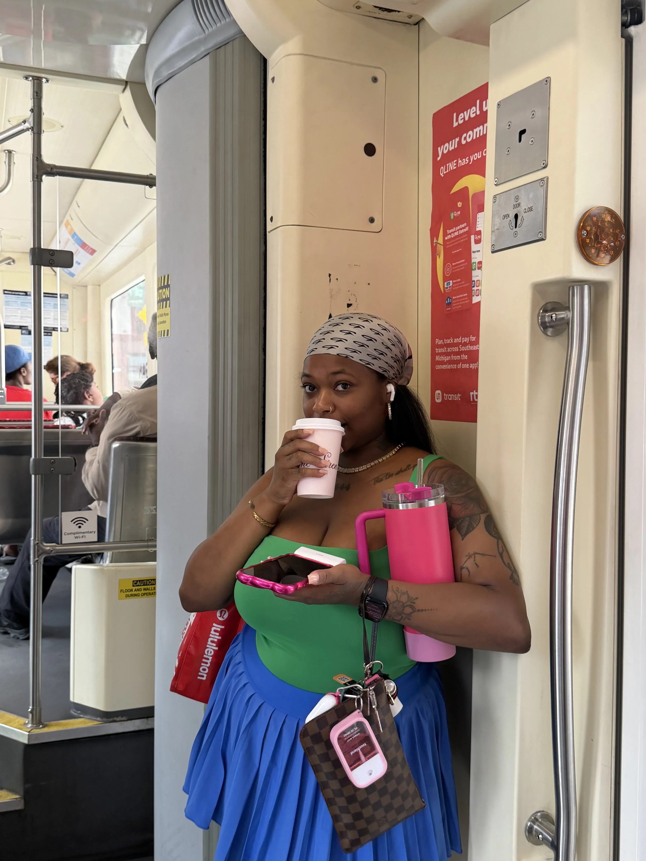 A woman wearing a green top and blue skirt drinking from a pink cup on a bus, with a brown checkered bag and various electronic accessories hanging from her waist. Passengers are visible in the background.