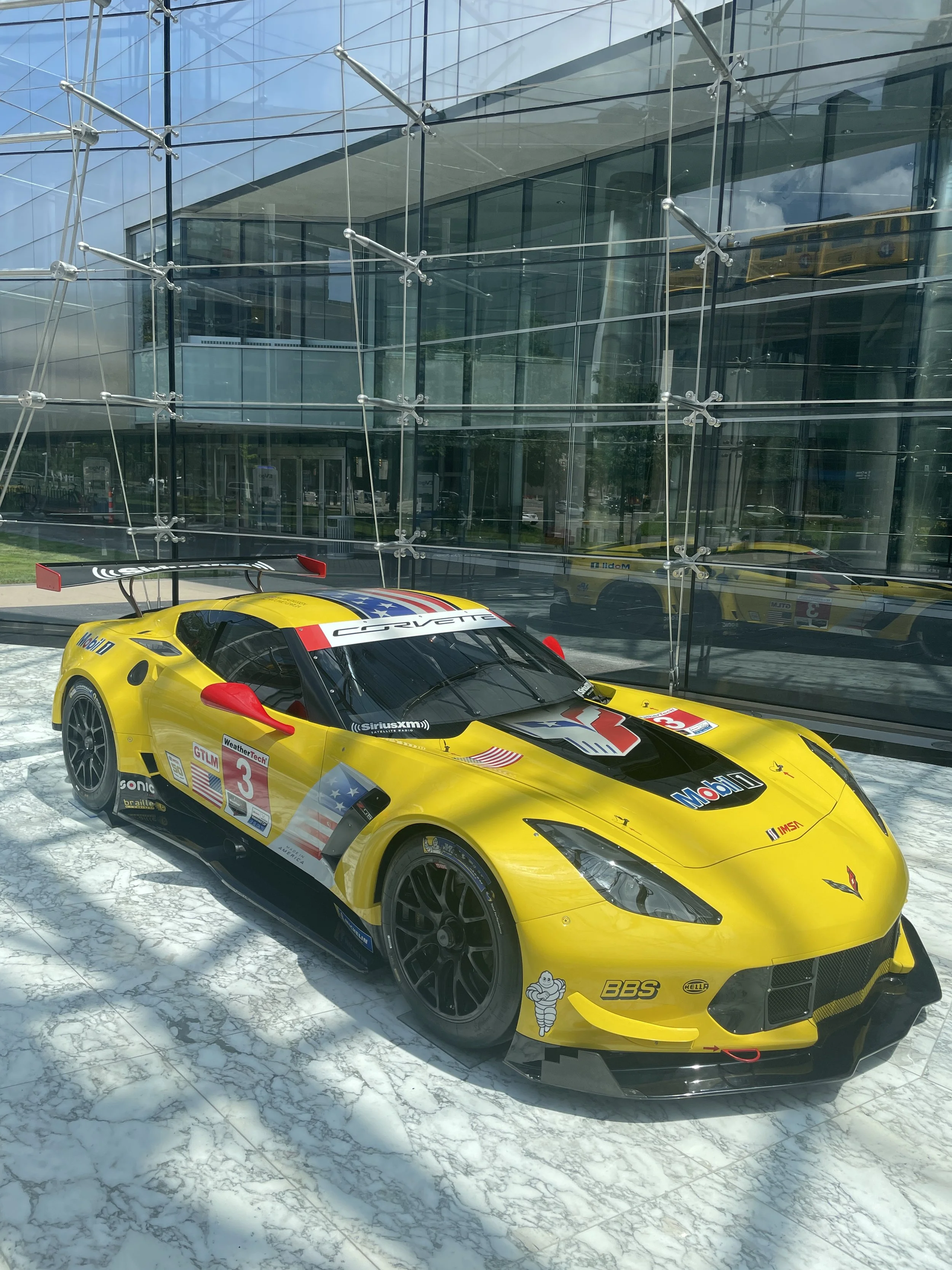 A yellow racing car with various sponsor logos, including Corvette and Michelin, parked indoors on a marble floor, with a modern glass building reflecting in the background.