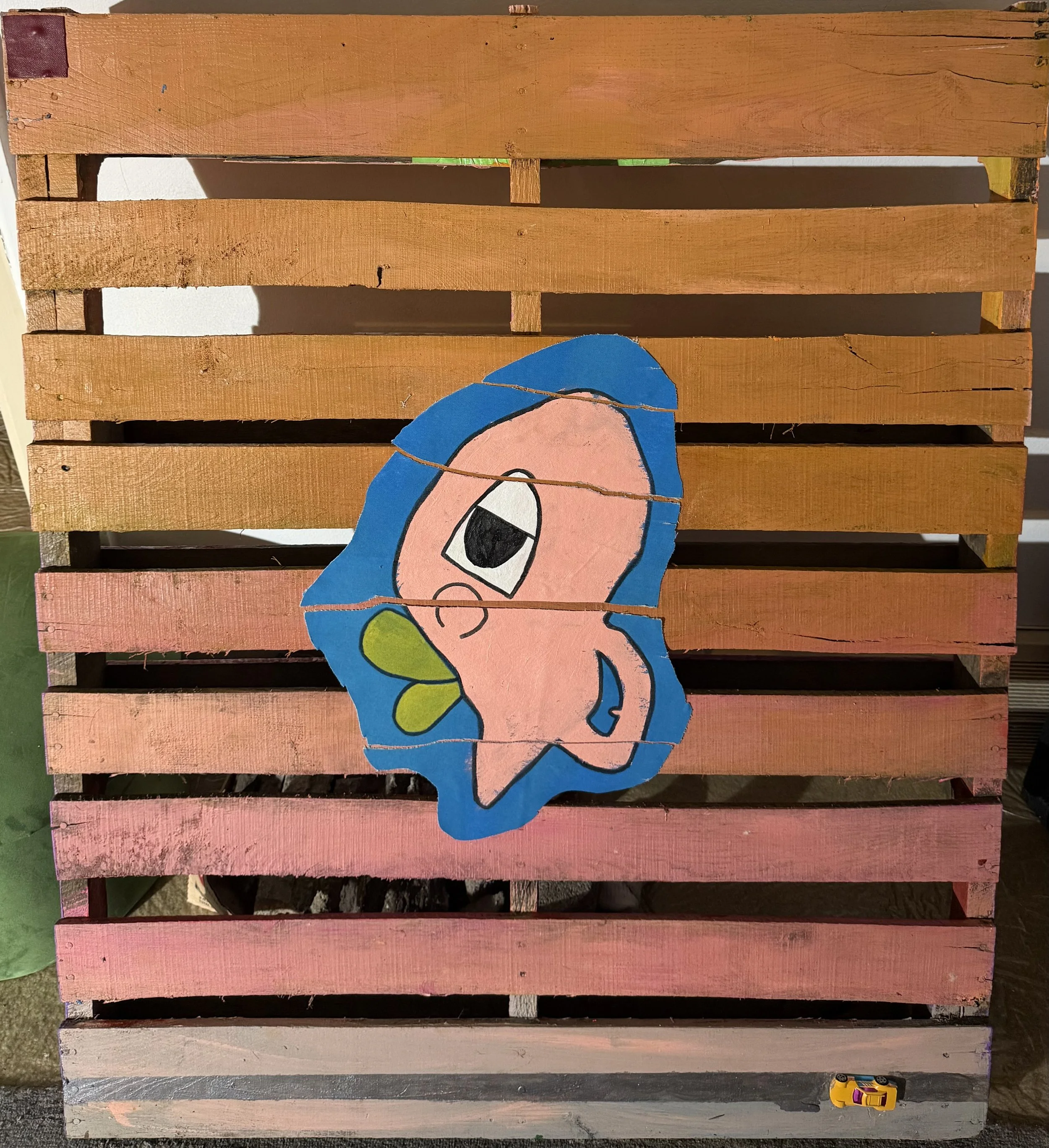 A colorful wooden pallet with painted cartoon face of a girl with blue hair and big eyes, and a small yellow toy car at the bottom right corner.