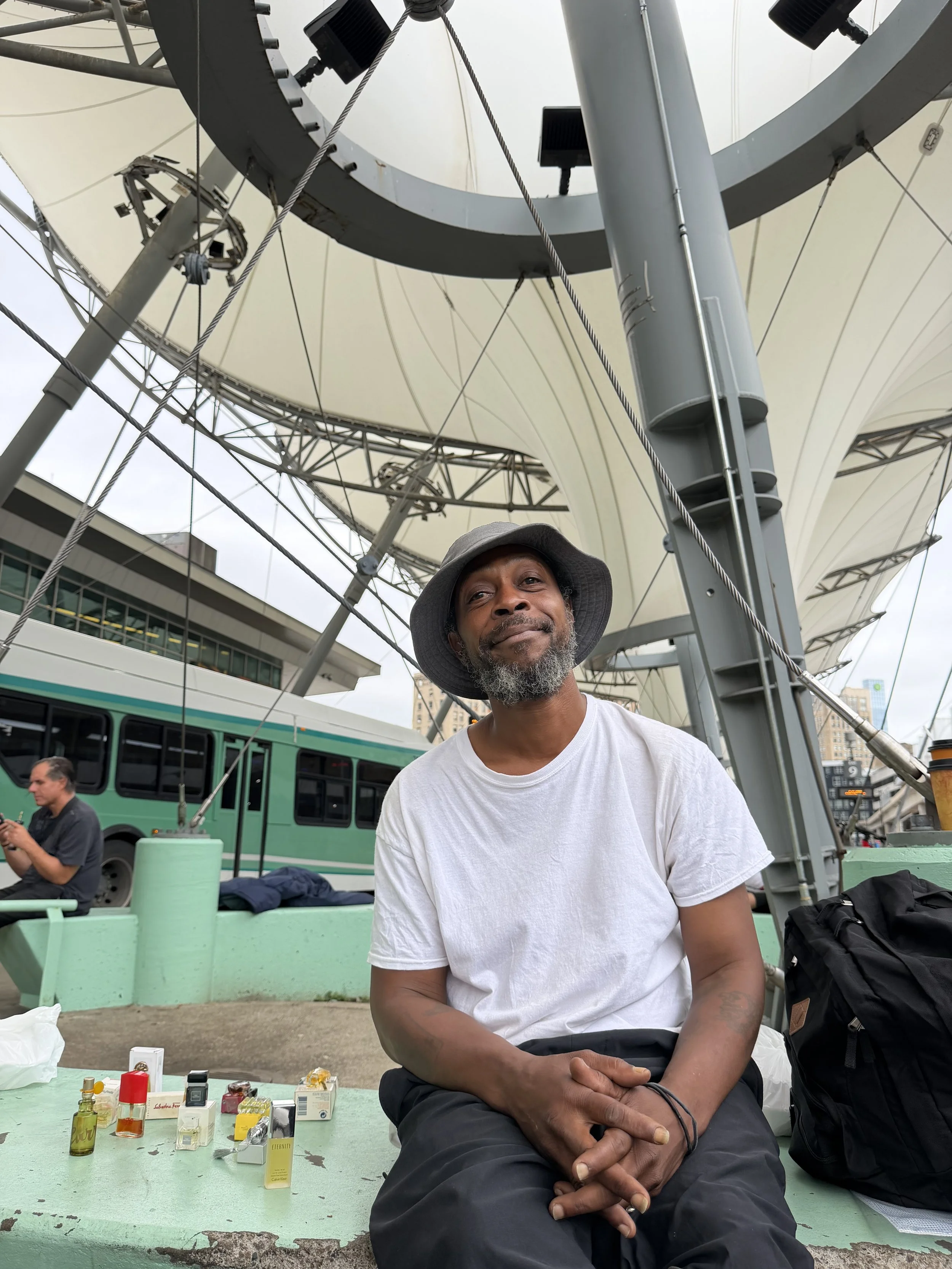 A man wearing a hat and a white T-shirt sitting on a green bench in an outdoor setting under a large parasol. There are perfume sample bottles on the table in front of him. In the background, a green bus and a person looking at their phone can be seen.