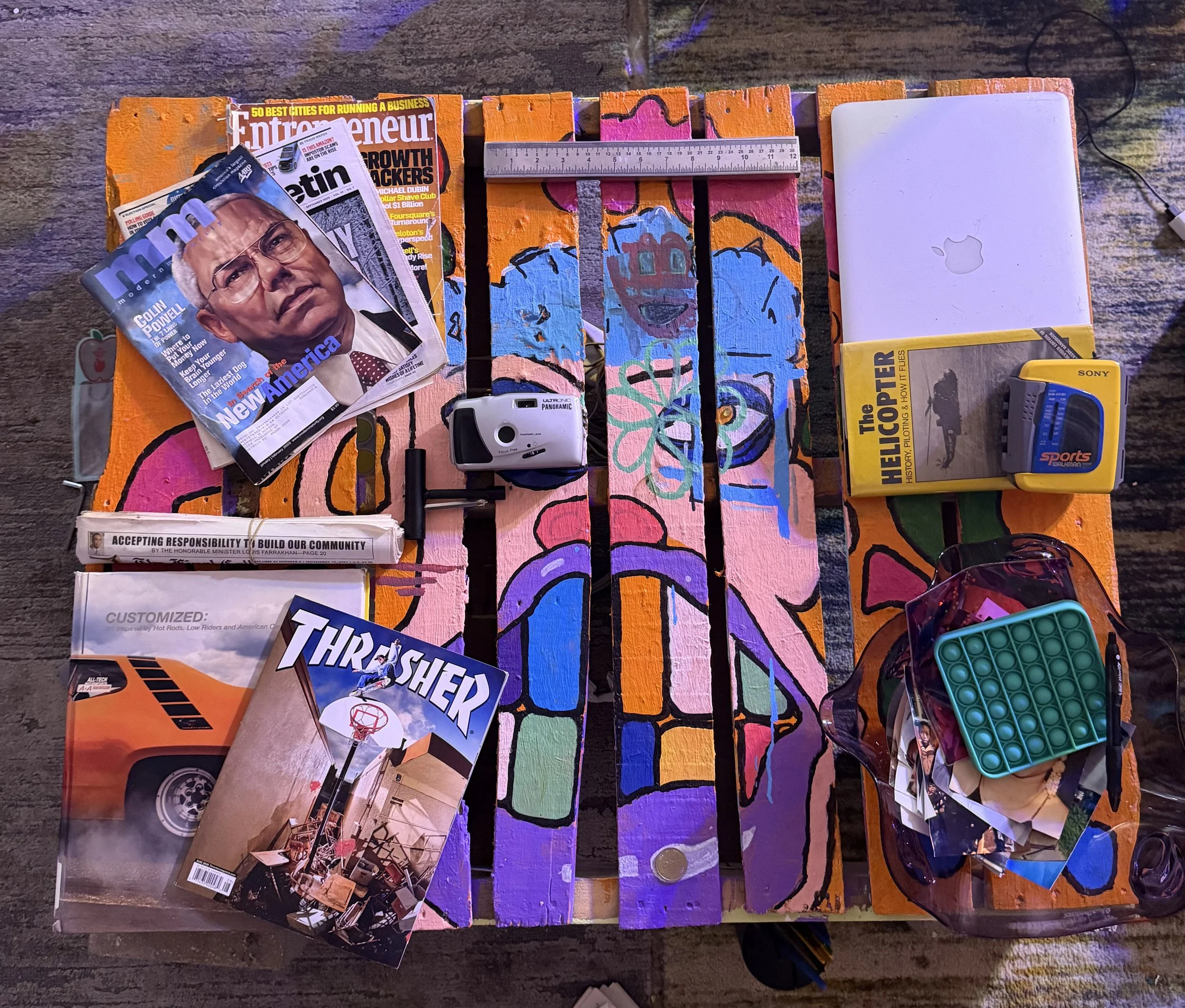 A colorful wooden art piece with various objects placed on top, including magazines, a ruler, a white electronic device, a yellow box, sunglasses, a container of slime, and photographs, all on a rough surface.