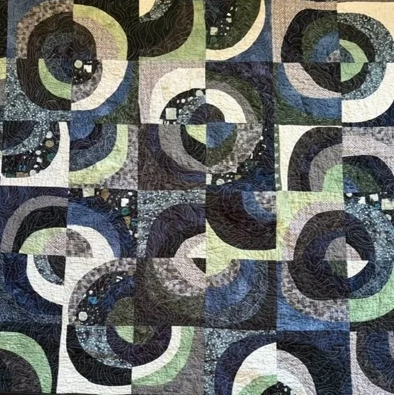 A close-up view of a quilt with a geometric, spiral pattern in dark and light colors, including black, navy, gray, white, and light green.