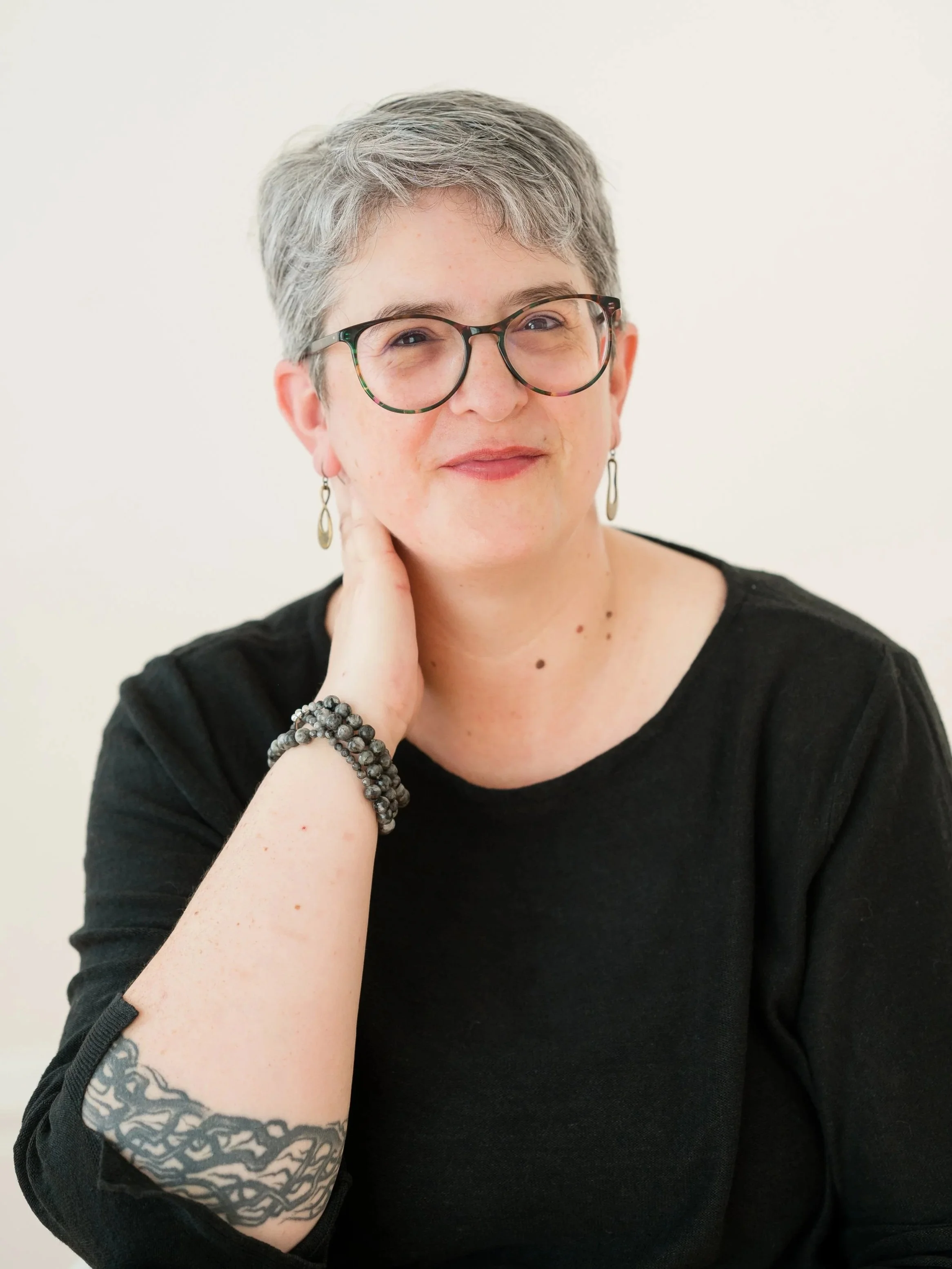 A woman with short gray hair, wearing glasses, earrings, and a black shirt, smiling and touching her neck, with a tattoo on her left forearm.