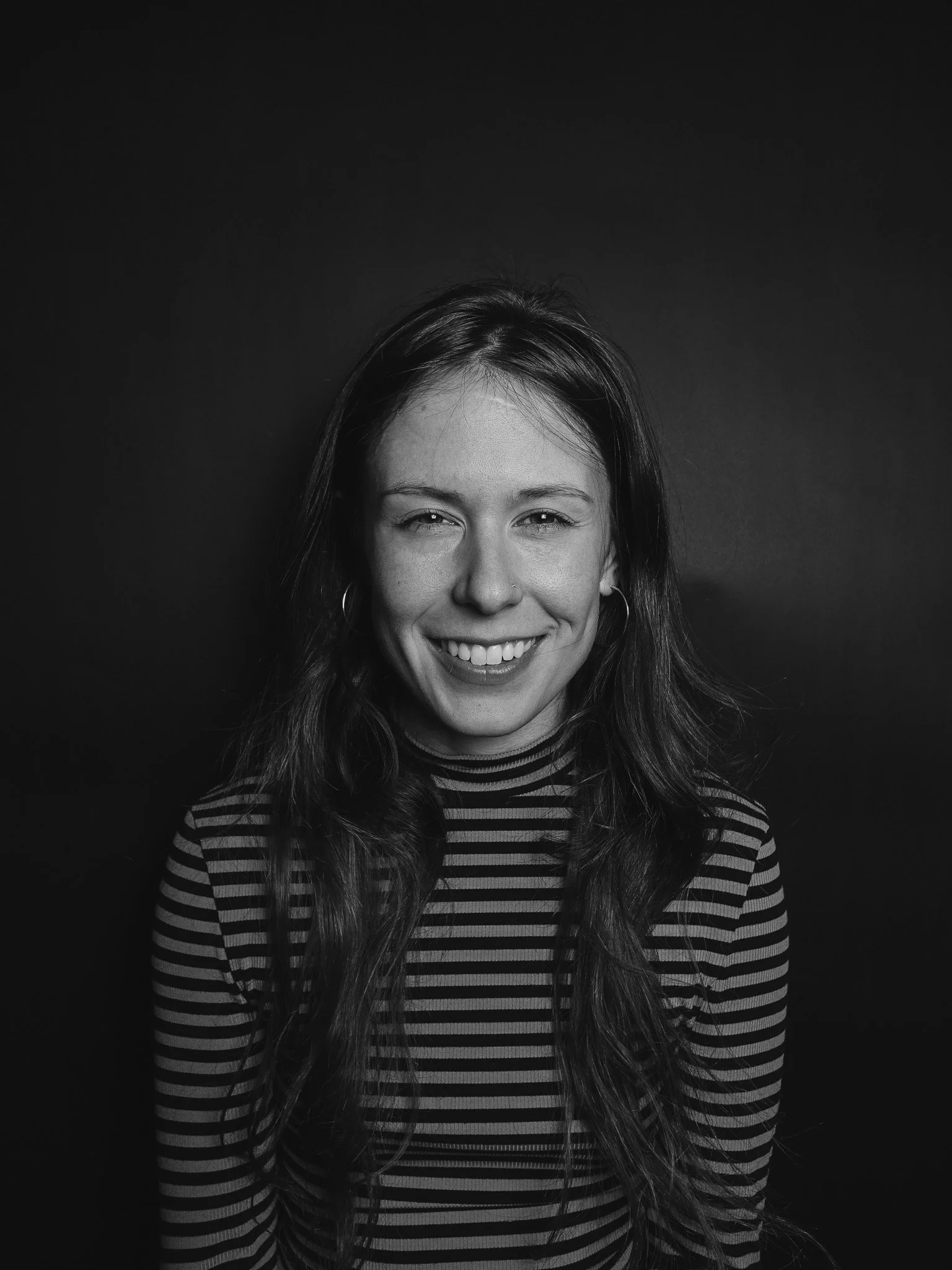 Black and white portrait of a smiling woman with long wavy hair wearing hoop earrings and a striped shirt, posing against a dark background.