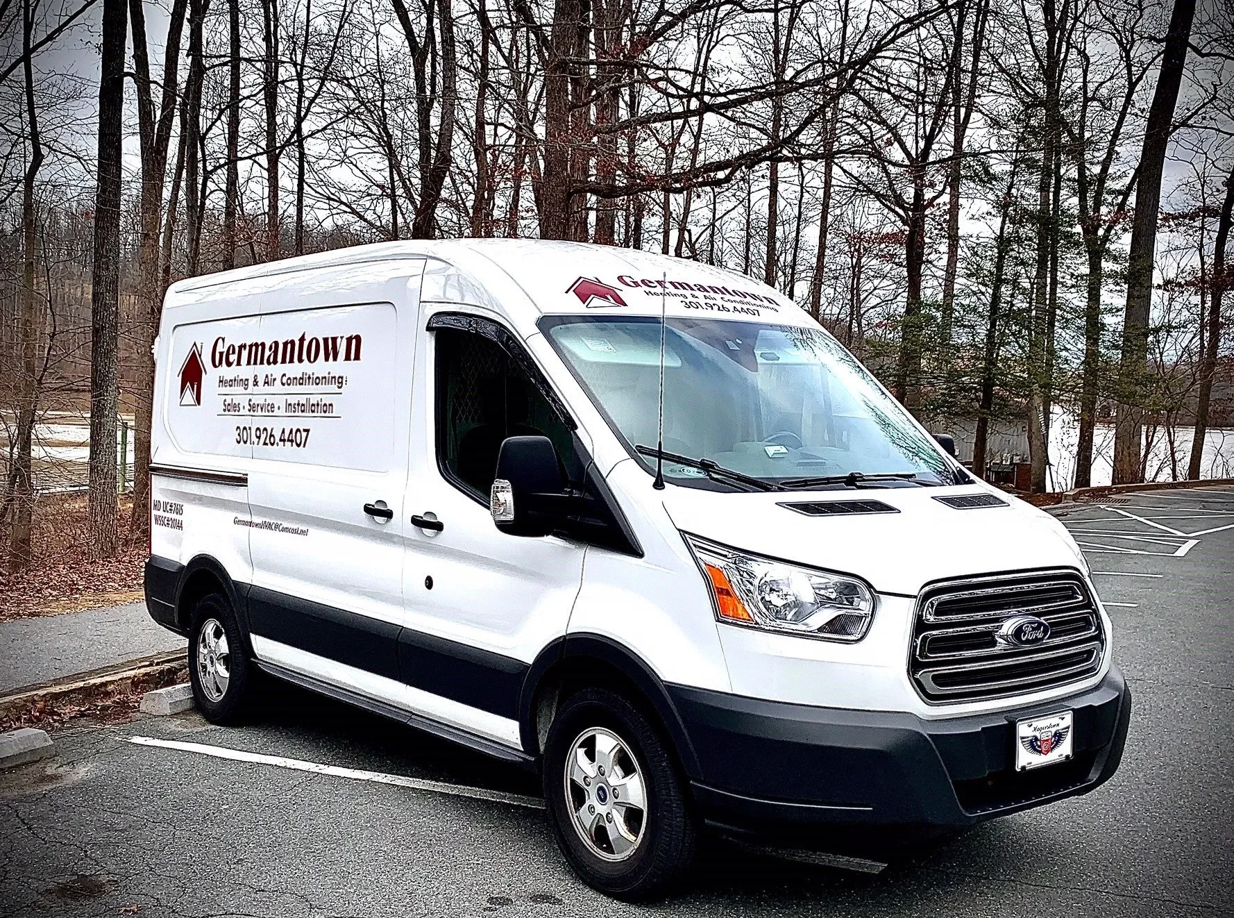A white commercial van parked in an outdoor parking lot surrounded by leafless trees. The van has branding on the side for Germantown Heating & Air Conditioning, with a logo, phone number 301.926.4407, and services listed as sales, service, and installation.