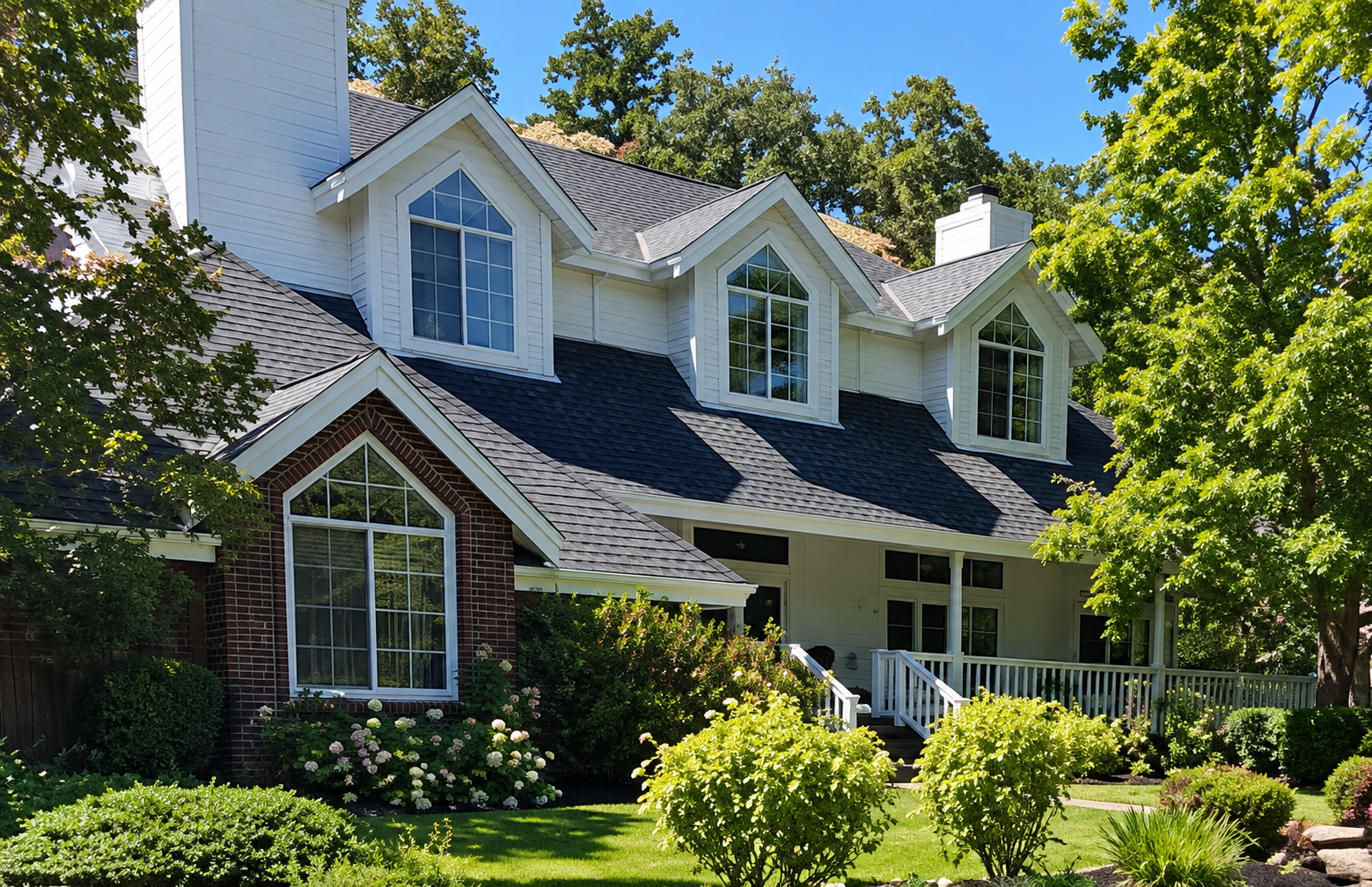 A two-story house with white siding and brick accents, multiple gabled roofs, large front windows, and a wooden front porch with white railings, surrounded by lush green trees and shrubs on a sunny day.