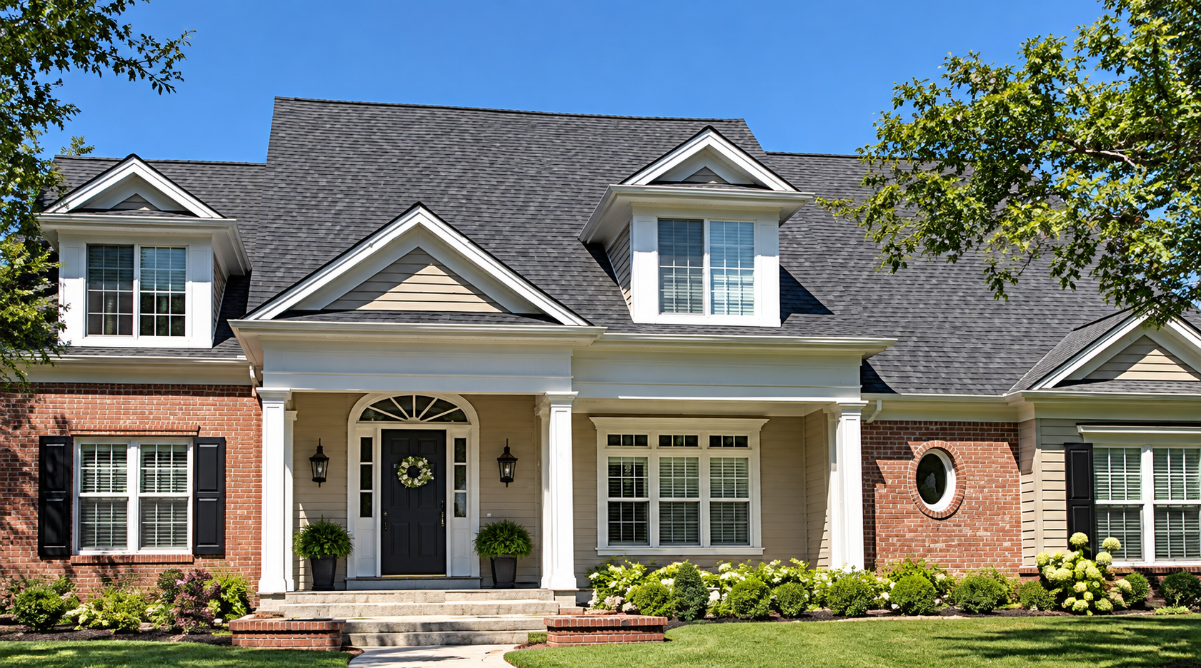 Front view of a house with a brick and siding exterior, black front door with wreath, gabled roof, and landscaped garden with bushes and flowers.