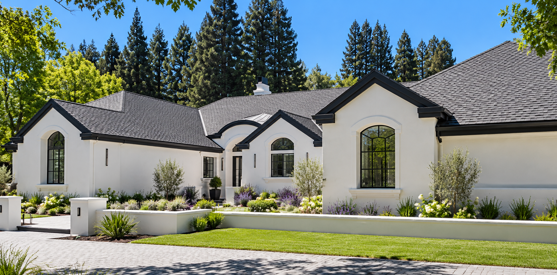 A modern white house with black window frames and a dark gray roof, surrounded by a landscaped garden with green grass, bushes, and small trees, under a clear blue sky.