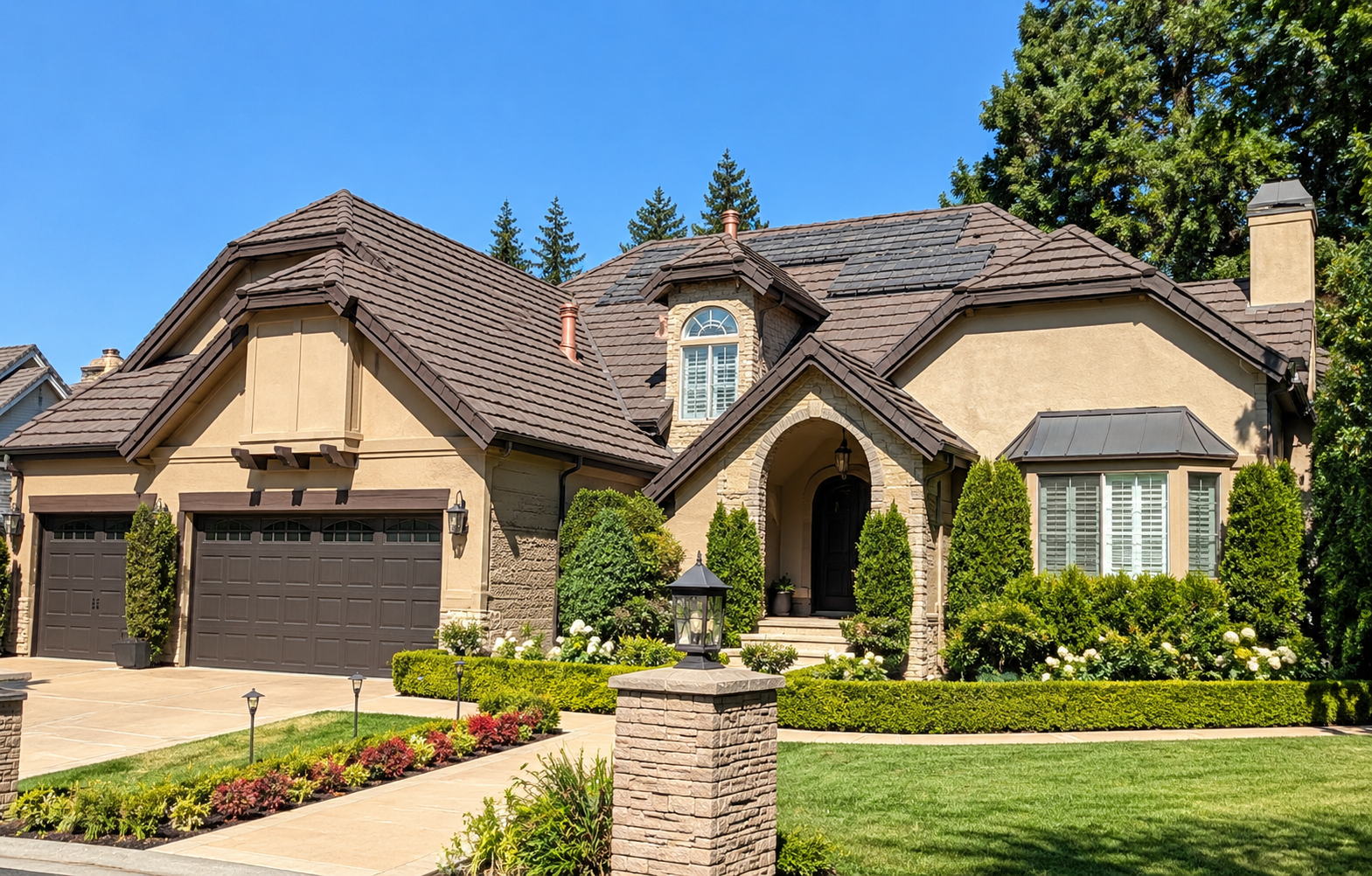 Large modern house with a manicured lawn, trees, and landscaped flower beds in front, beige exterior walls, dark garage doors, and a tiled roof.