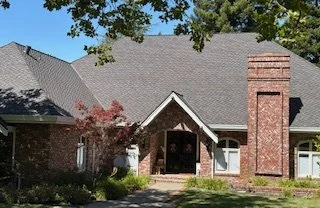 Front view of a brick house with a gabled roof, entrance porch, and tall chimney, surrounded by trees and landscaping.