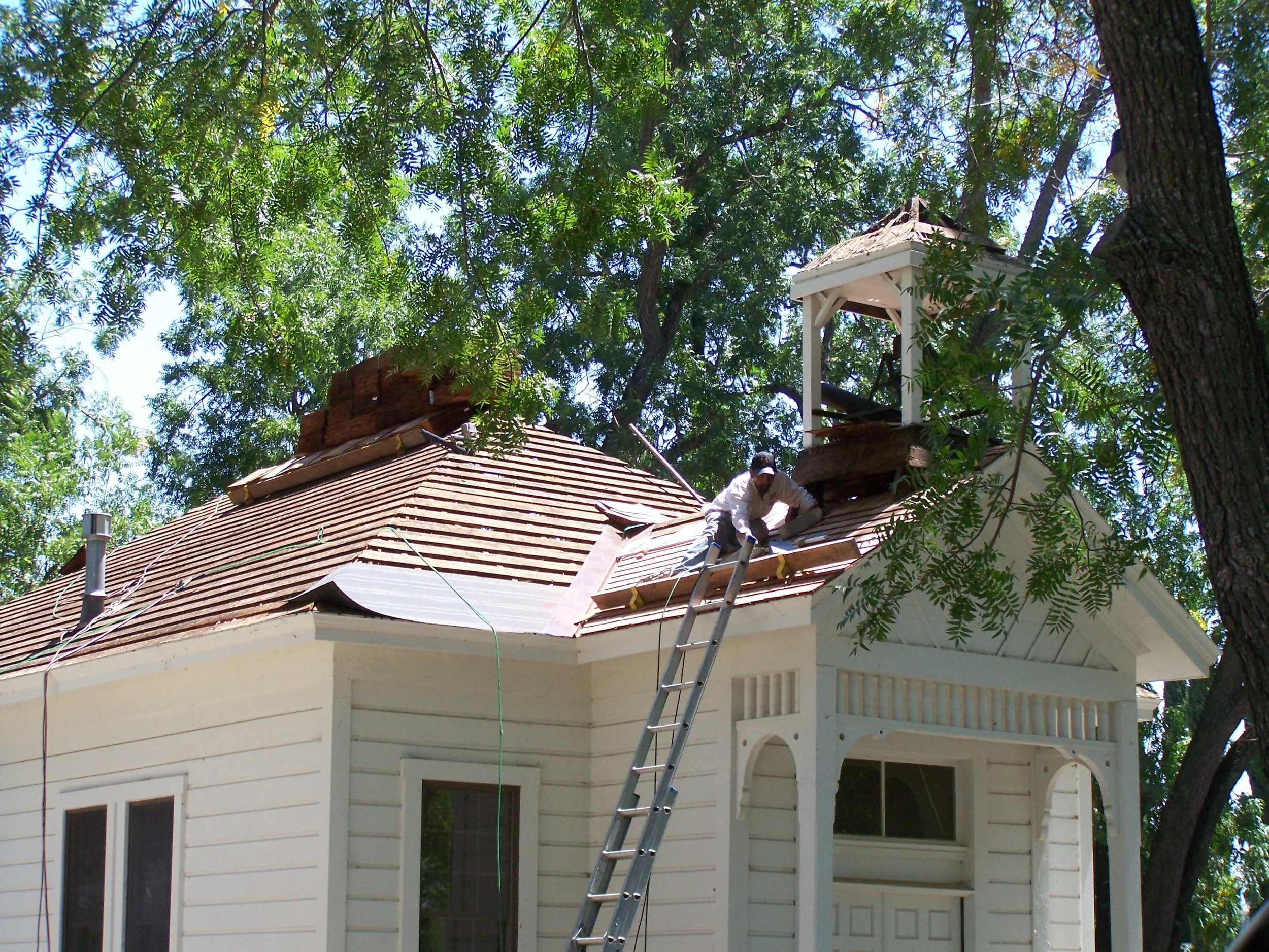 A house with a brown tiled roof under construction, with a person working on the roof, surrounded by tall trees and green foliage.