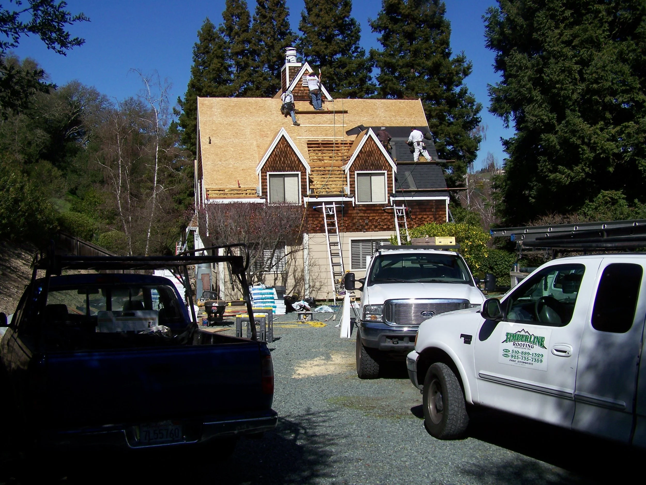 Construction workers are installing new roofing on a house with a steep roof. Several vehicles, including trucks and cars, are parked in the foreground, and tools and materials are scattered around the site.