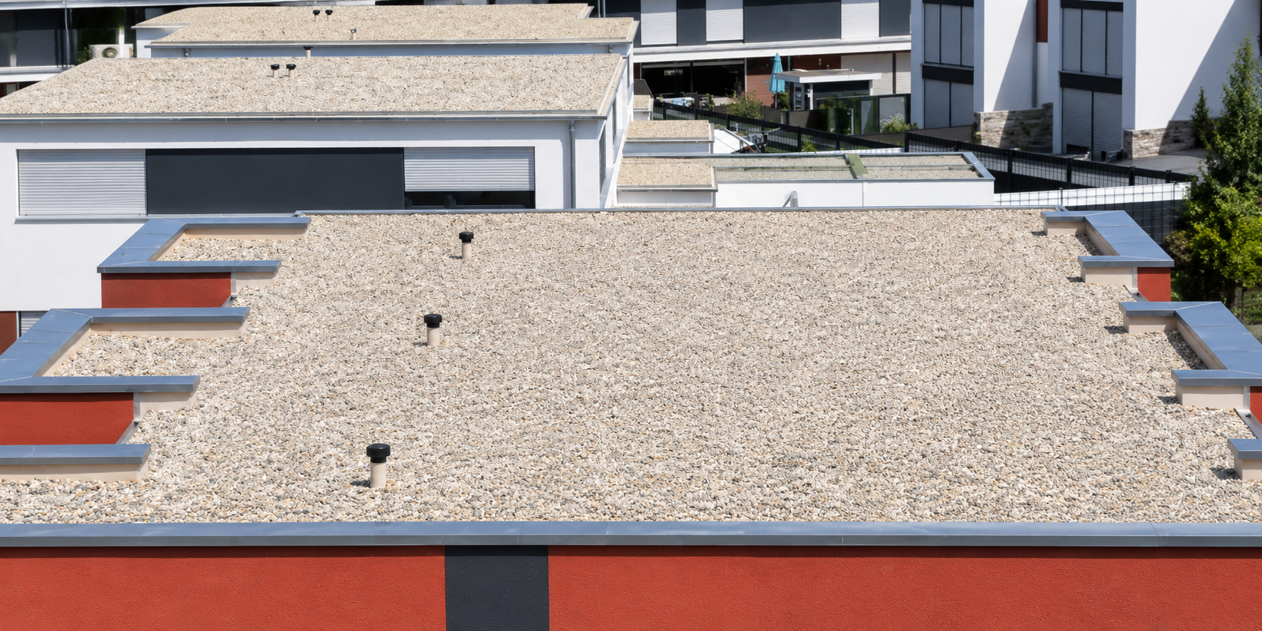 View of multiple modern apartment rooftops with gravel and metal accents, surrounded by residential buildings and greenery.