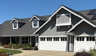 A large suburban house with a dark gray roof, white garage doors, and a covered front porch.