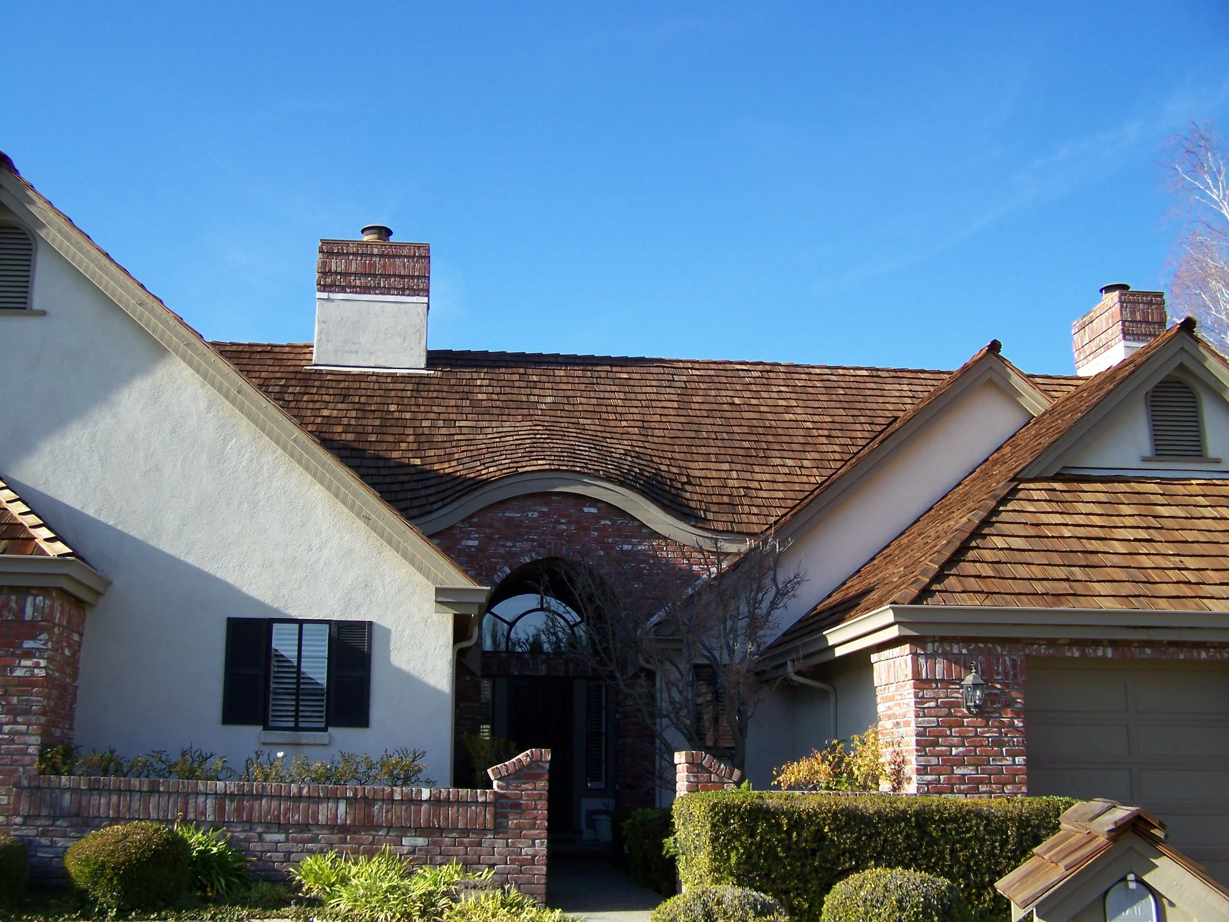Front view of a house with a sloped tiled roof, brick chimneys, white walls, black shutters, and well-maintained shrubs in the yard.
