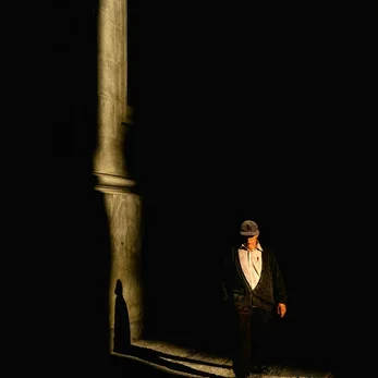 A person standing in a shadowy alleyway, lit by a single light source, with a tall wall on one side.