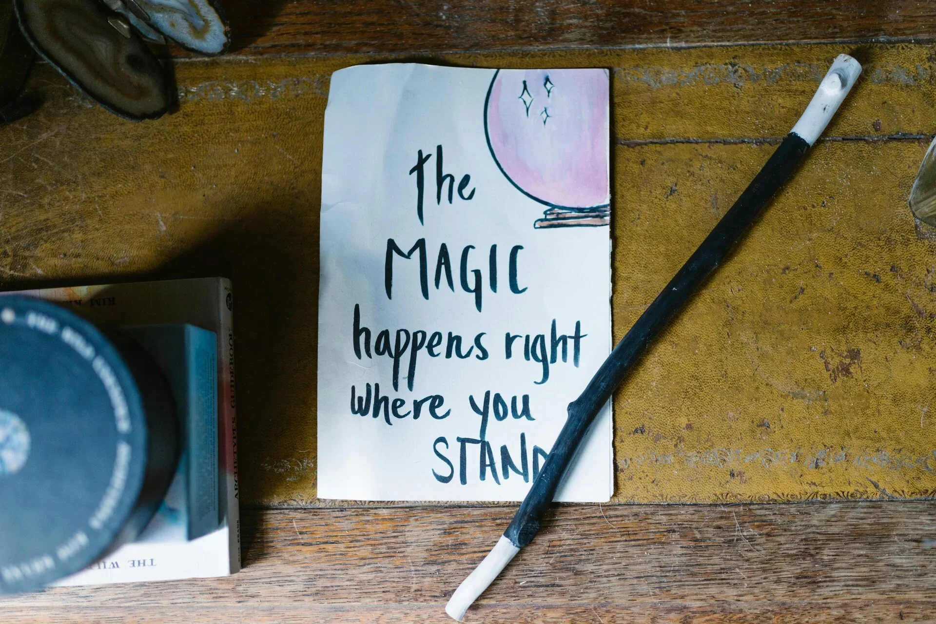A handwritten sign on a piece of paper that says, 'the magic happens right where you stand.' The note is placed on a wooden surface next to a black and white cigarette and a partially visible camera lens. There are other objects on the table, including some books or boxes and a glass.