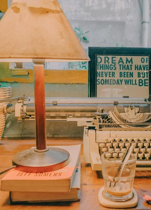 A vintage desk scene with a typewriter, a glass of beverage with a straw, a lamp, a book, and a framed quote that says, "Dream of things that have never been but someday will be."