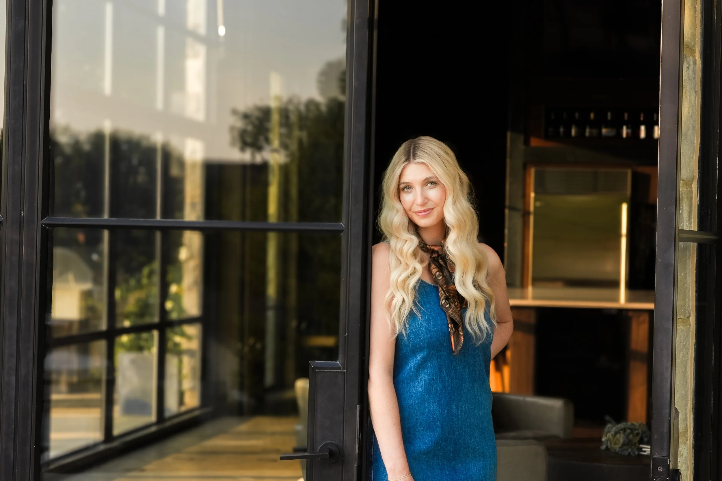 A woman with long, wavy blonde hair standing in a doorway, wearing a blue dress and looking at the camera.