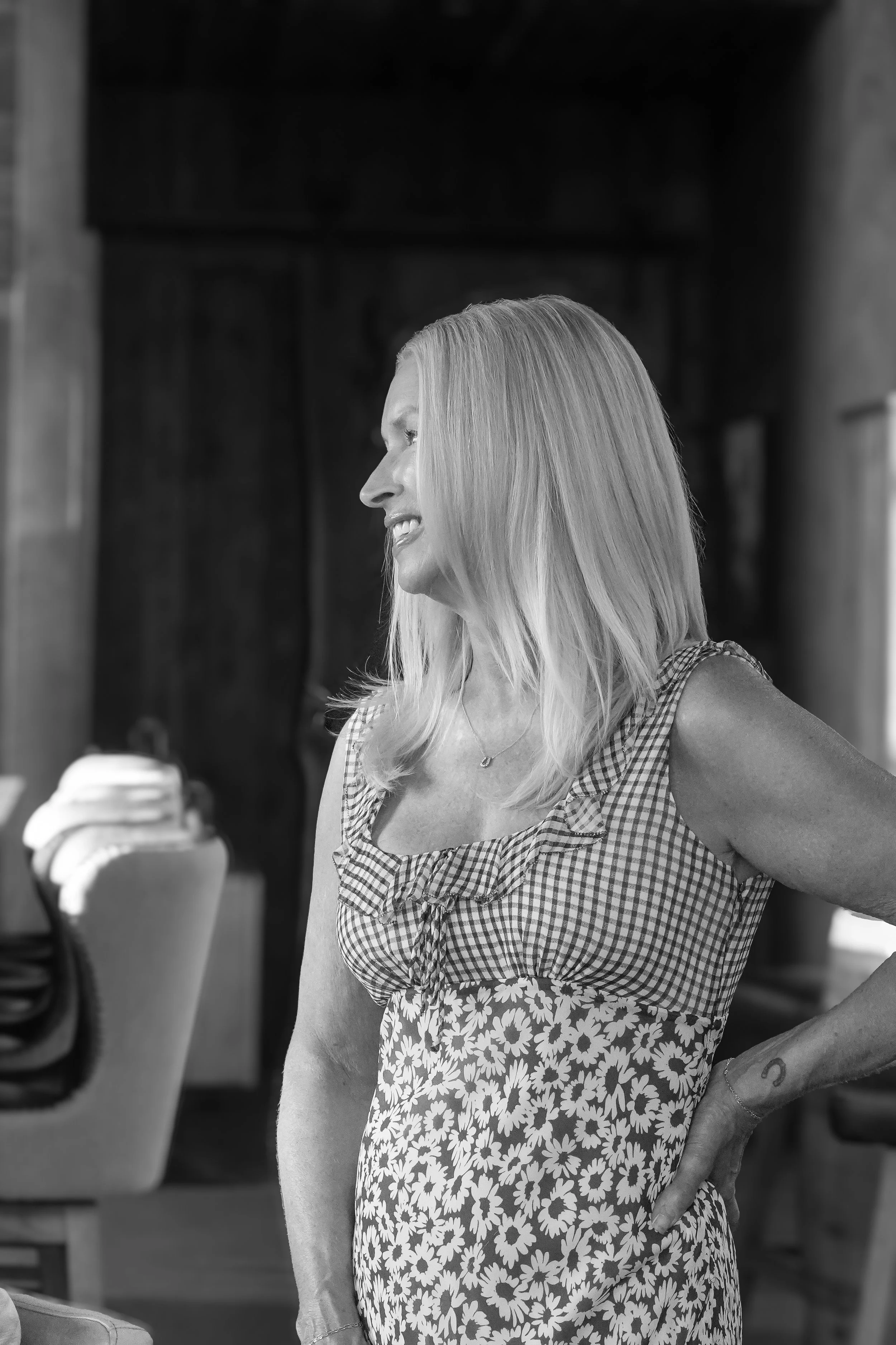 A black and white photo of a woman with long, straight hair, smiling and wearing a sleeveless, gingham top with ruffle details and a floral skirt. She is standing indoors, with visible chairs and blurred background.