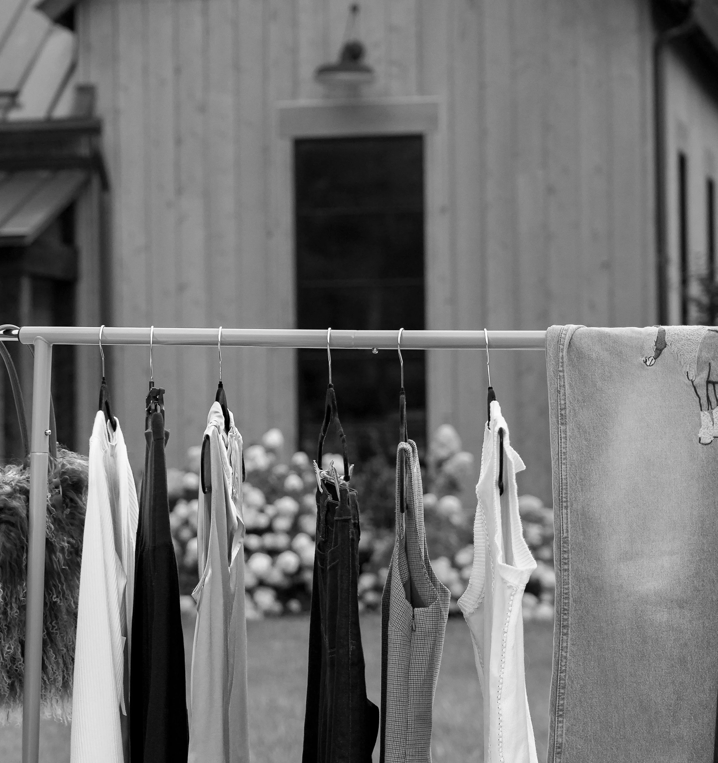 Clothes hanging on a clothing rack outside, with a wood-paneled wall and some blurred objects or flowers in the background.