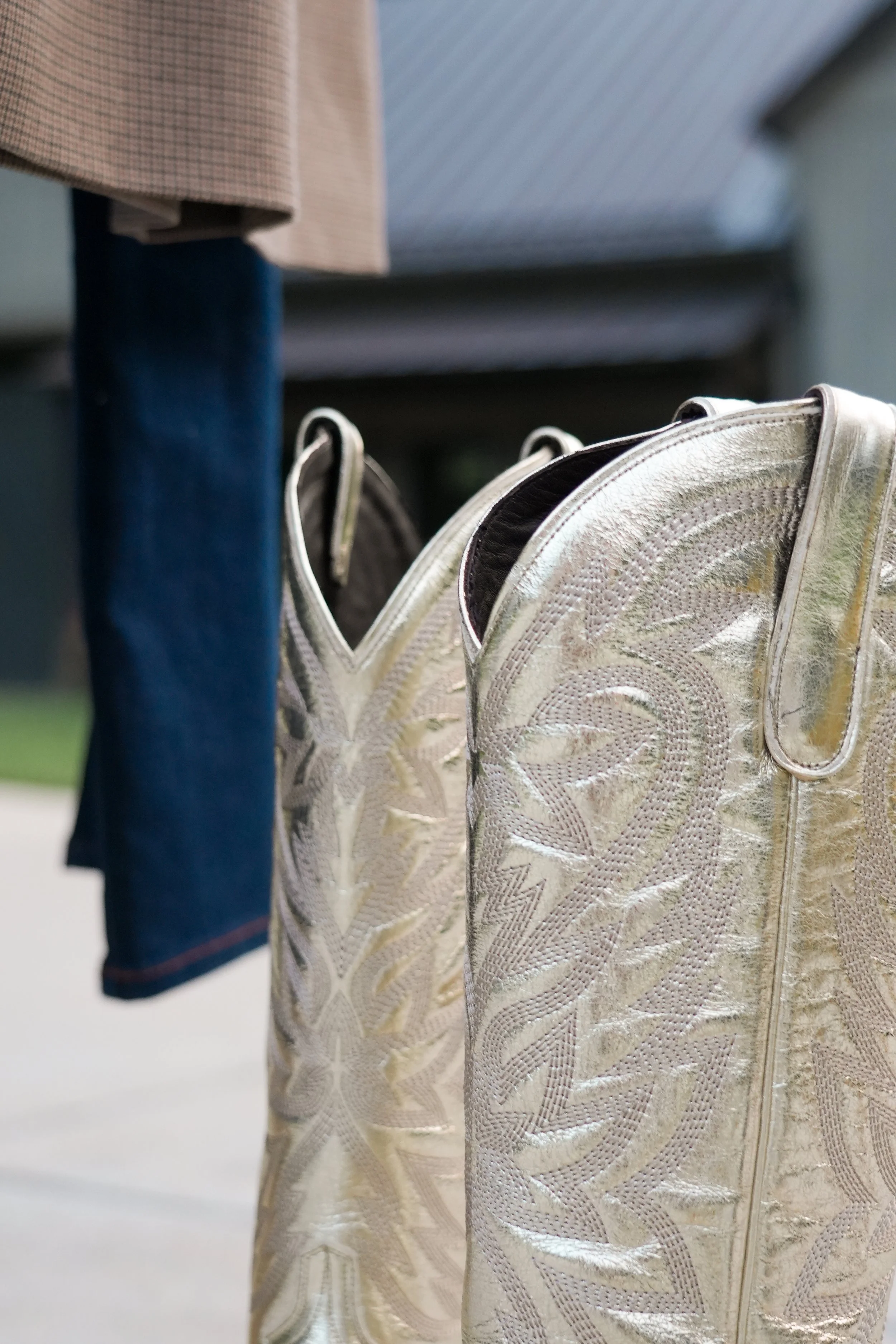 A pair of metallic, shiny cowboy boots with intricate patterns, with a plaid shirt and blue jeans also hanging nearby.