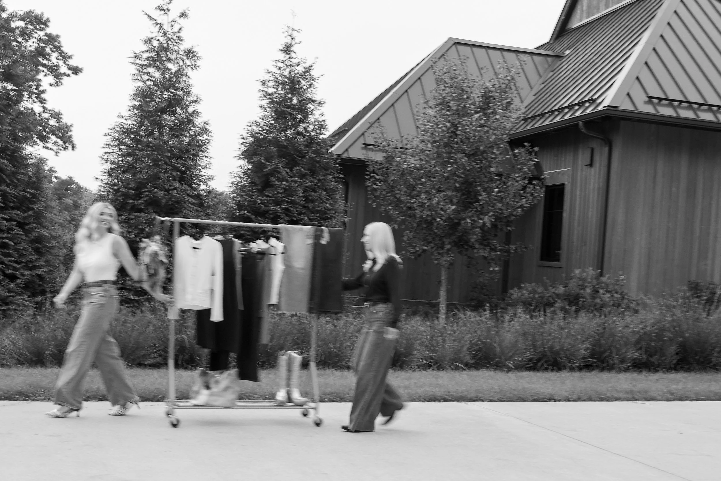 People walking on a sidewalk near a house with trees in the yard, a clothing rack with clothes hanging on it, and a small tree in the outdoor space.