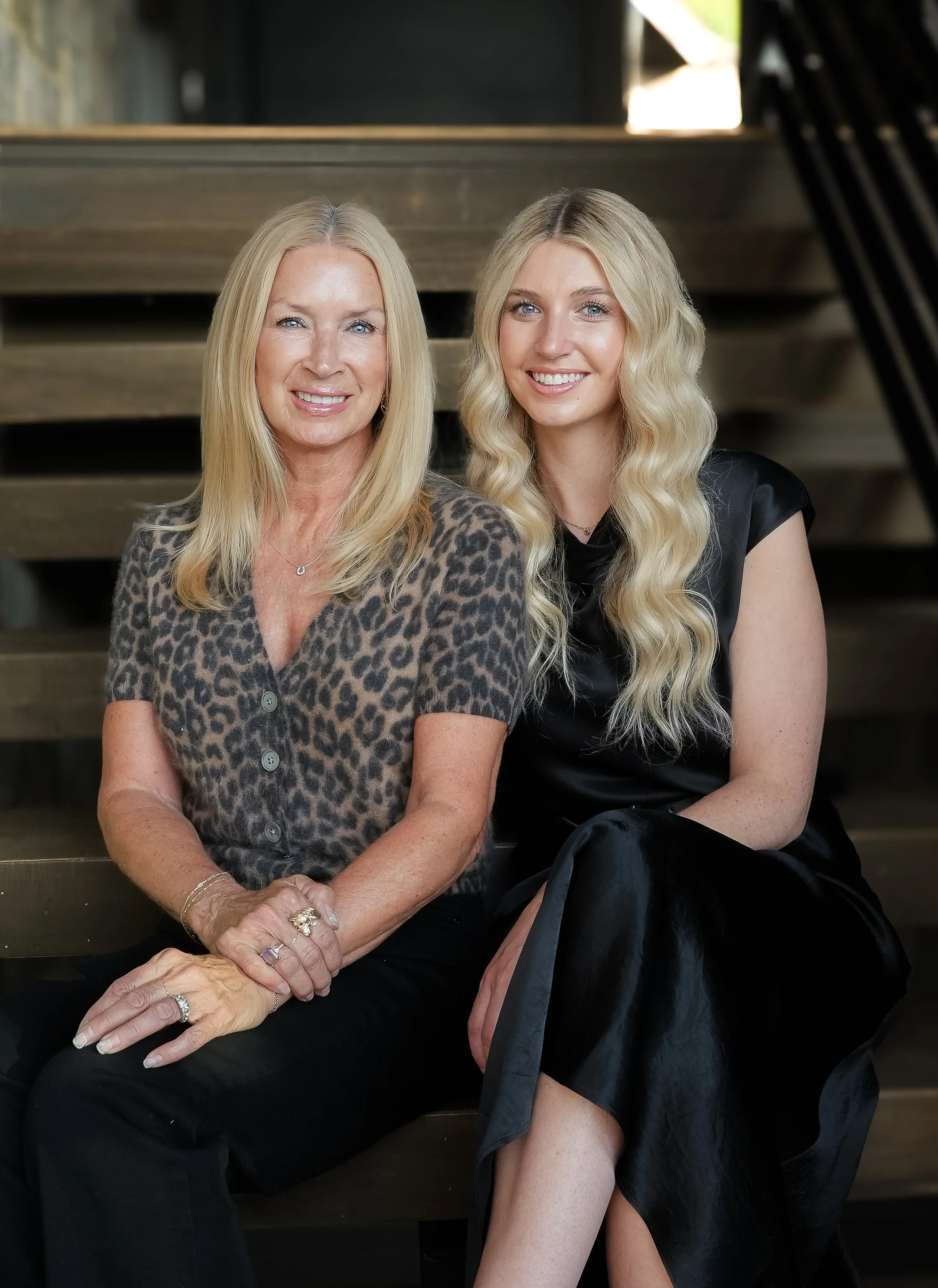 Two women sitting on stairs, smiling at the camera, with natural light coming from behind, one wearing a leopard print top and the other wearing a black satin dress with open cardigan.