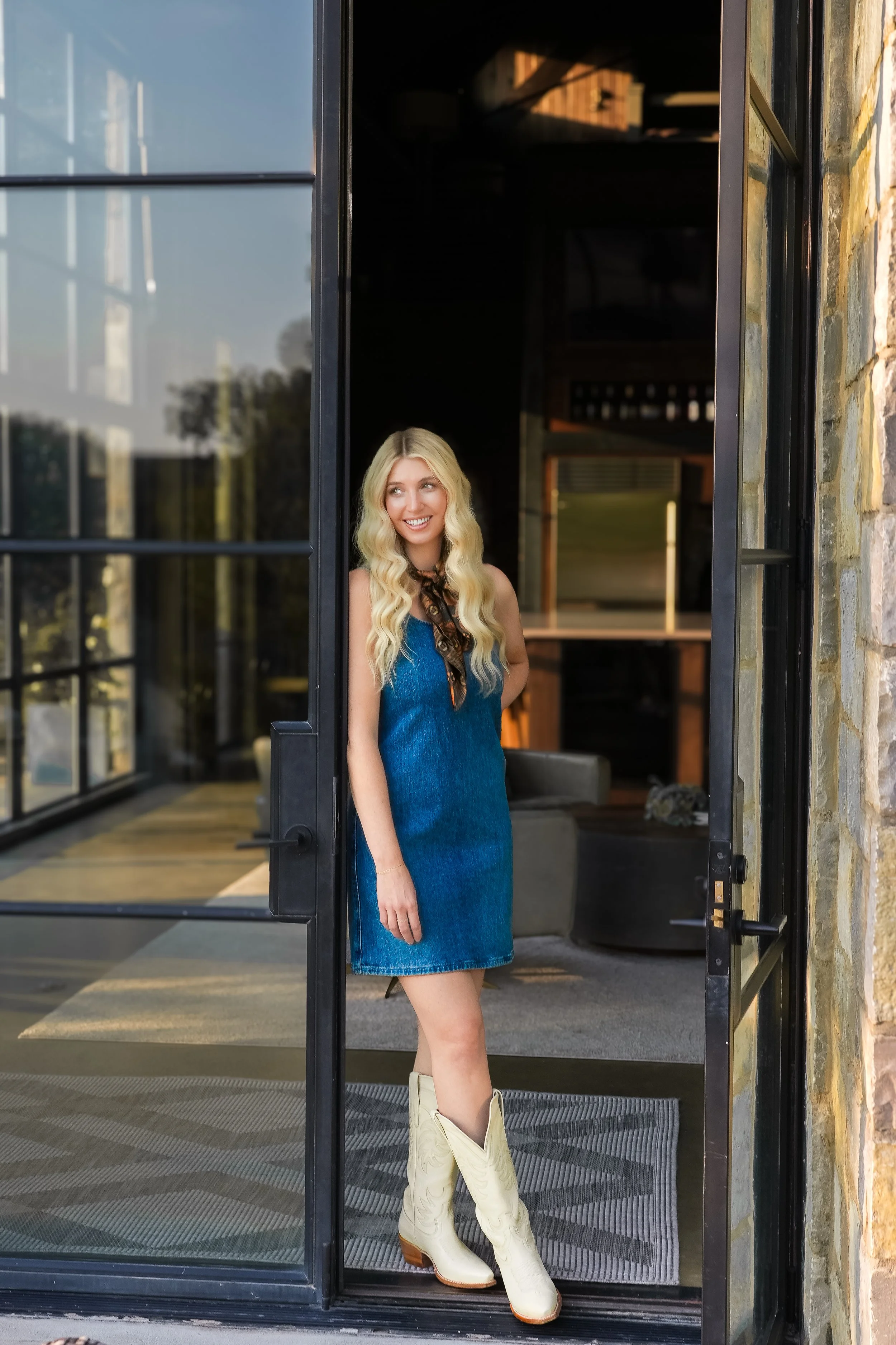 A woman with long blonde hair wearing a blue dress and white cowboy boots, standing and smiling at an open glass door.