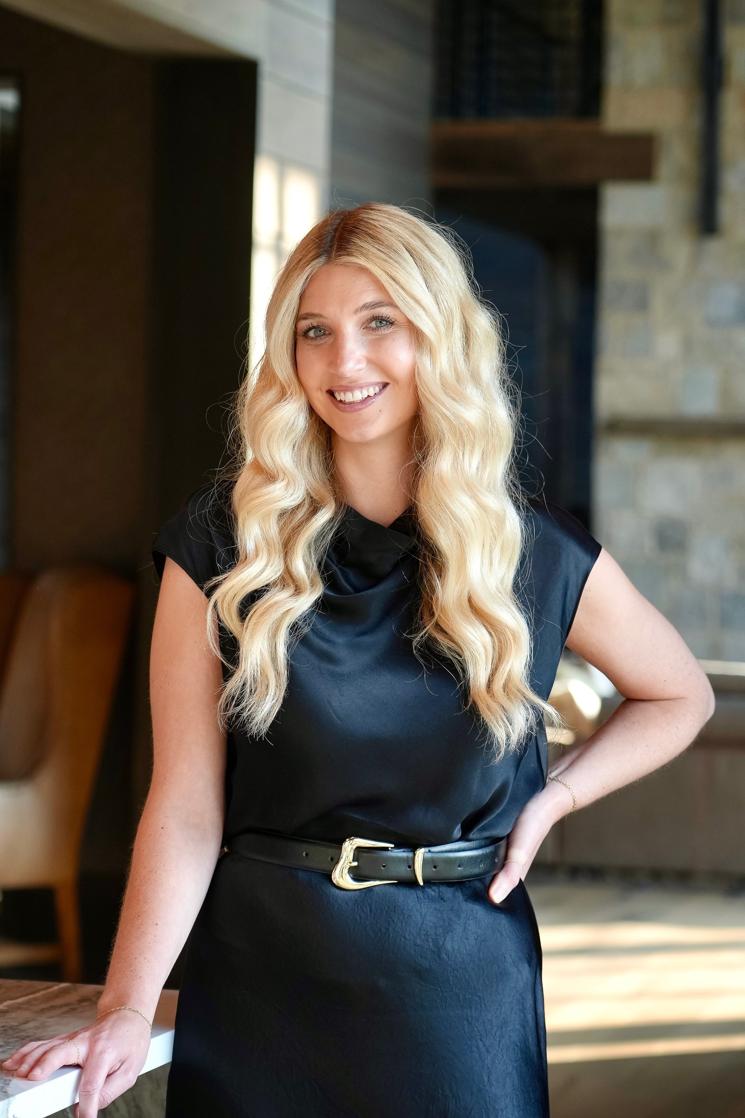 A woman with long blonde wavy hair, wearing a black dress with a belt, leaning with her left arm on a white countertop inside a modern indoor space.