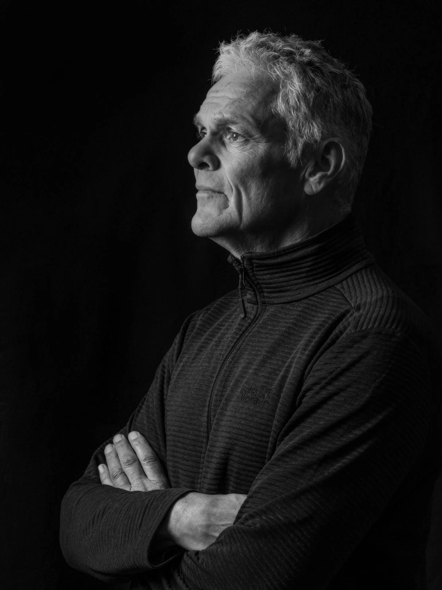 Zwart-wit portret van een oudere man met grijs haar, gekleed in een jas, met armen over elkaar, kijkend naar rechts, minimale achtergrond.