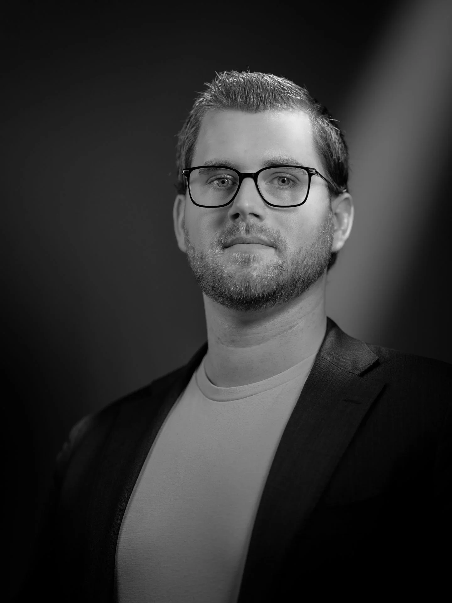 Zwart-wit foto van een man met bril, sportief kapsel, lichte huid en een lichte baard, gekleed in een zwarte blazer en een wit shirt, kijkend naar de camera met een neutrale uitdrukking.