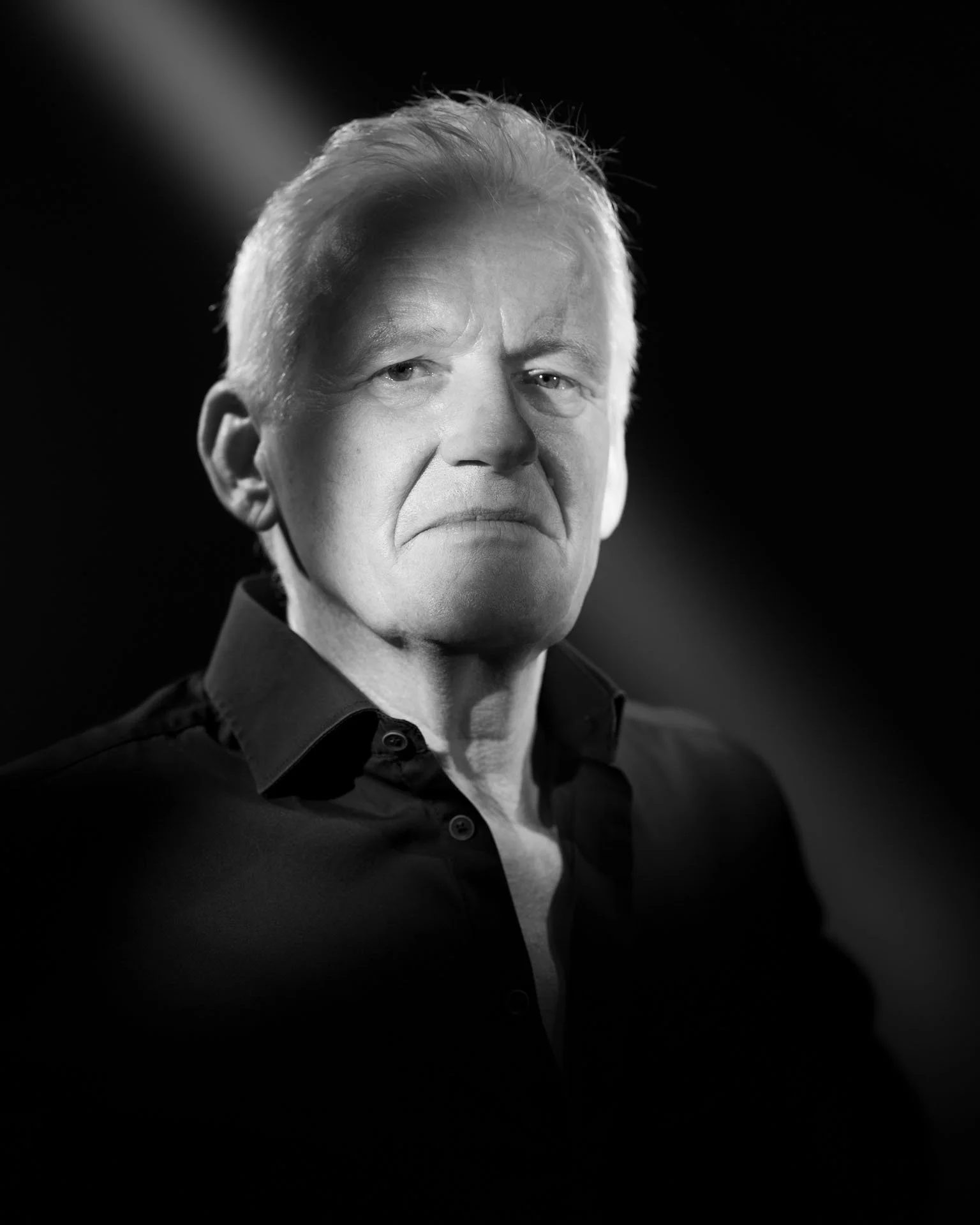 Zwart-wit foto van een man met steekhoudende uitdrukking, donkere achtergrond, gekleed in een zwart overhemd.