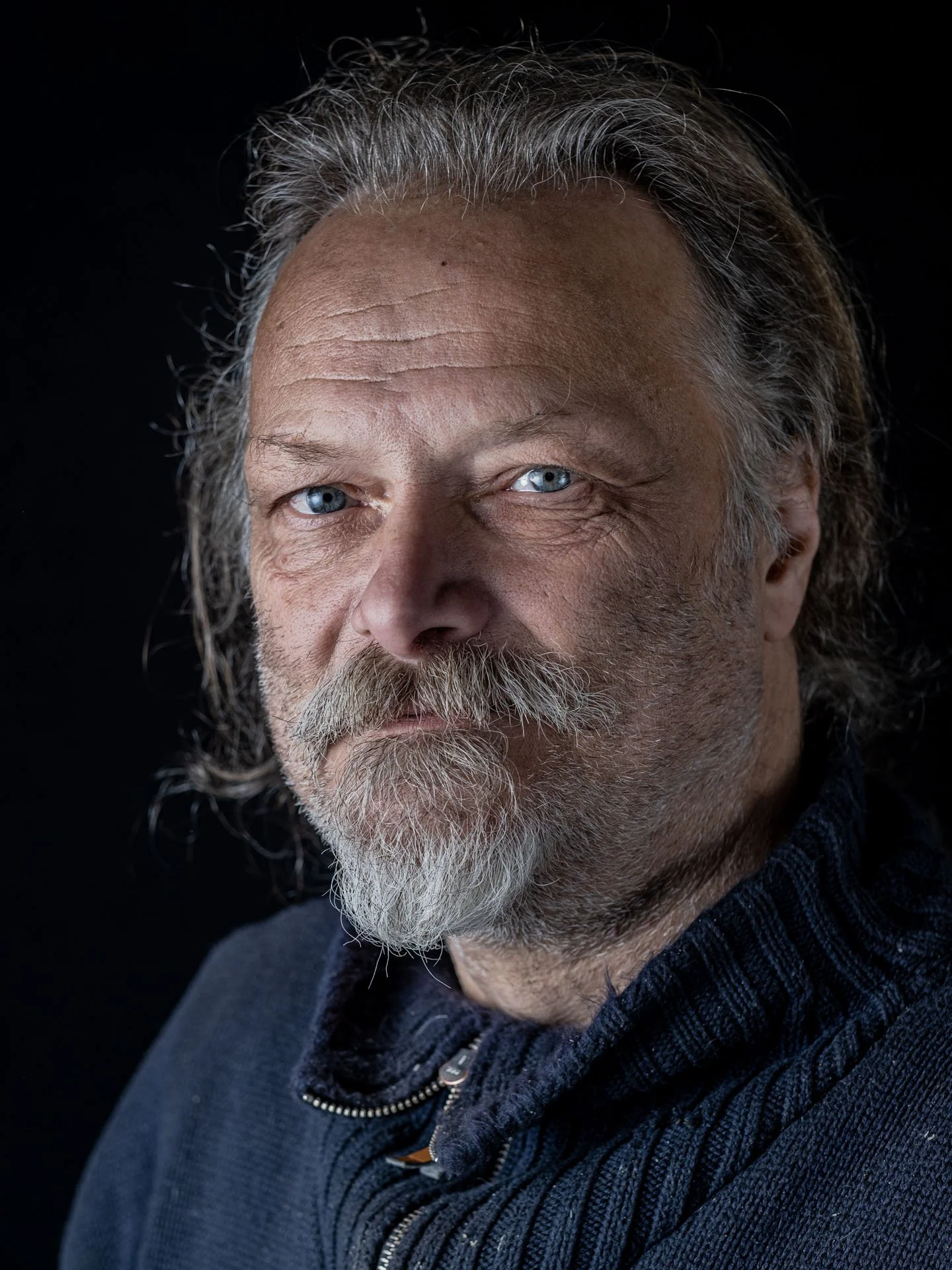 Close-up van een oudere man met grijs haar en een volle baard, gekleed in een donkerblauwe sweater, tegen een zwarte achtergrond.
