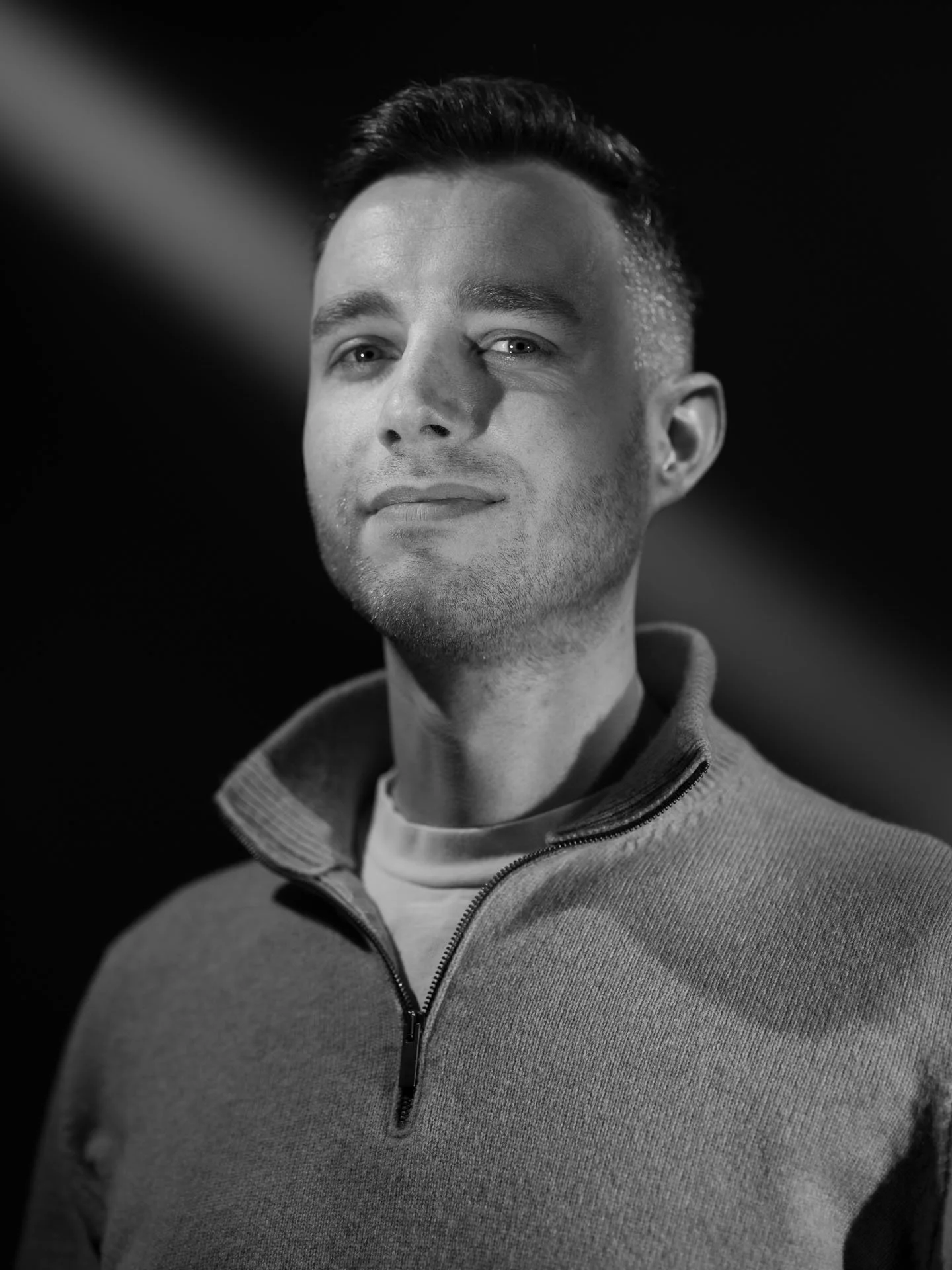 Portret van een jonge man met korte, donkere haar en lichte ogen, gekleed in een grijs zip-up sweater en een wit shirt, zwart-wit foto