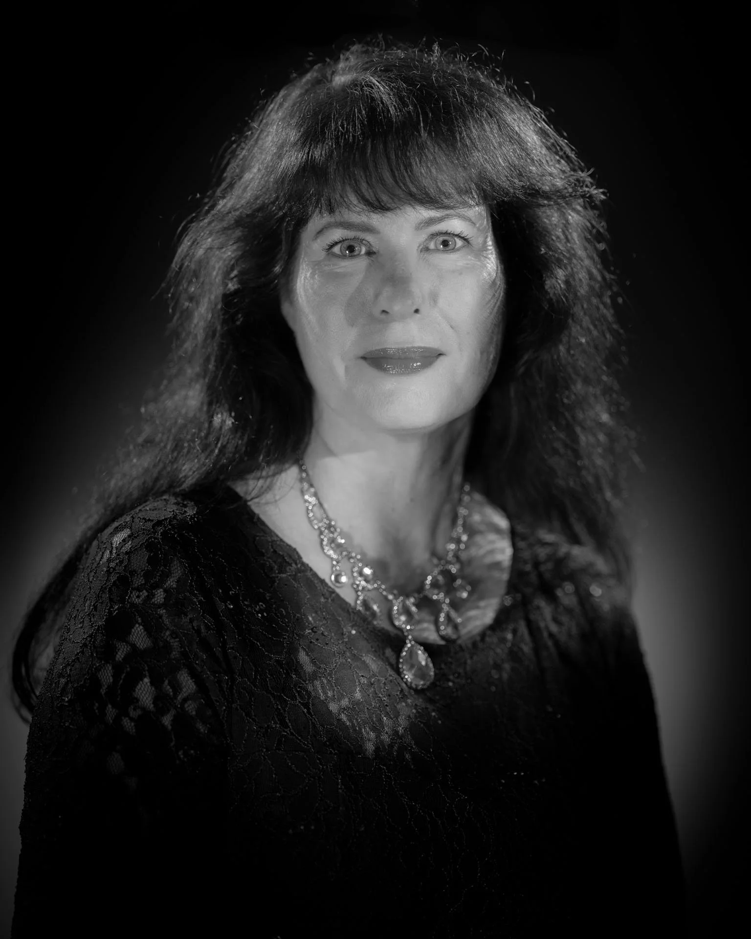Zwart-wit portretfoto van een vrouw met donkere, golvende haar, dragend een kanten jurk en een sierlijke ketting.